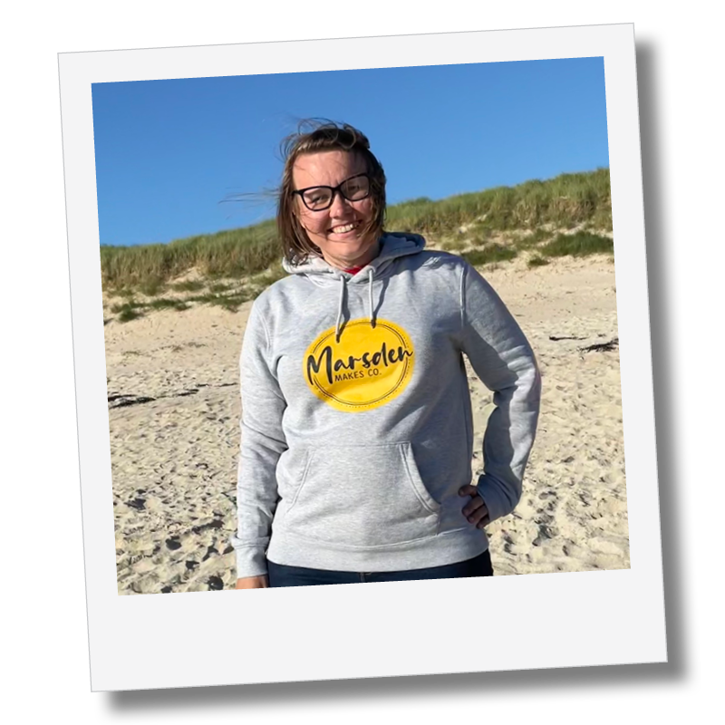 A woman standing on a sandy beach with grassy dunes in the background, wearing glasses and a gray hoodie with a yellow logo that reads 'Marsten Makes Co.', smiling at the camera.