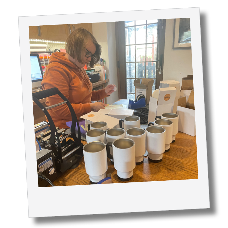 Marsden Makes Co Core Value Quality A woman with glasses and a brown hoodie standing at a cluttered wooden table, packaging or preparing items among multiple white cups, with boxes and a computer monitor in the background, near a glass door.