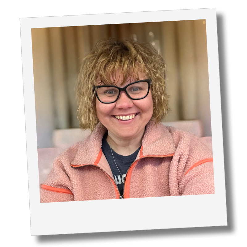 A woman with curly blonde hair, black glasses, and a pink zip-up fleece smiling at the camera indoors.