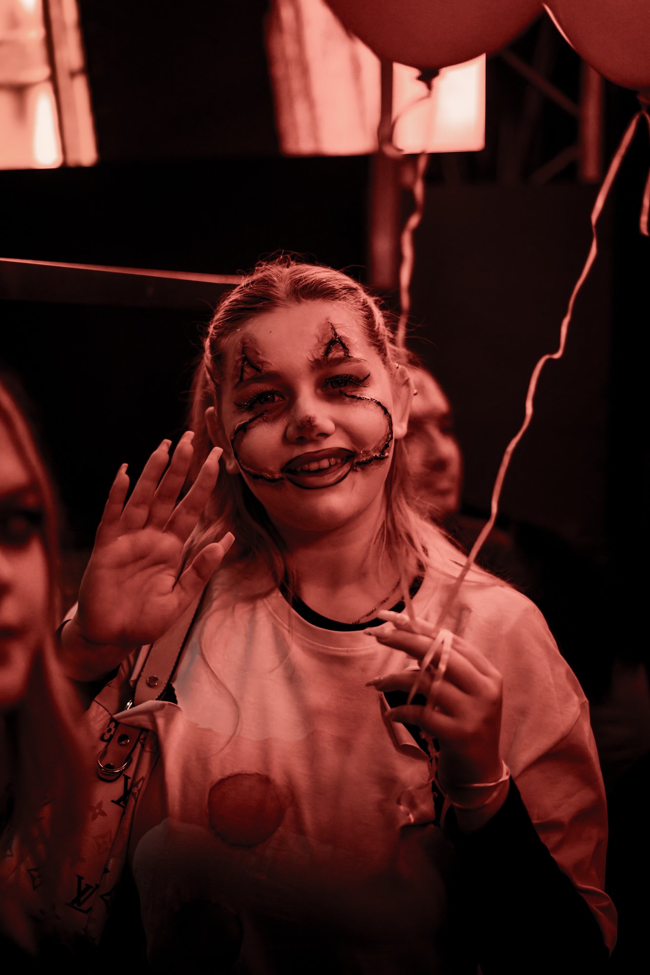 Junge Frau mit schrägem Make-up und lachender Miene bei einer Party, hält einen Ballon. Die Beleuchtung ist rot. Sie winkt zur Kamera.