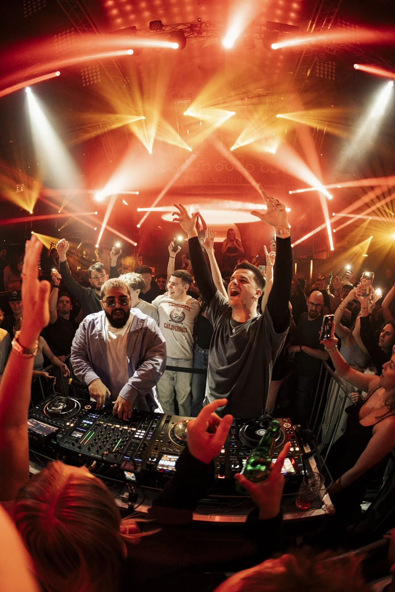 Menschen tanzen und feiern bei einem DJ-Set in einem Club mit rotem und orangefarbenem Licht und Disco-Effekt.