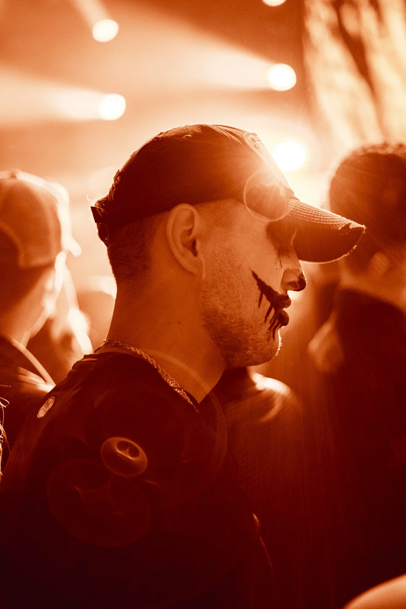 Ein Mann mit einem Baseballcap trägt clownähnliches Make-up und eine Clownnase, umgeben von anderen Personen in einem warmroten, beleuchteten Raum.