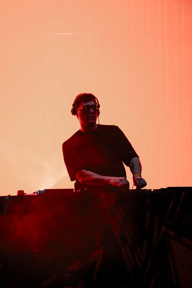 DJ mit Kopfhörern und dunkler Kleidung hinter einem DJ-Controller vor einem orange-roten Hintergrund bei einem Musik-Event.