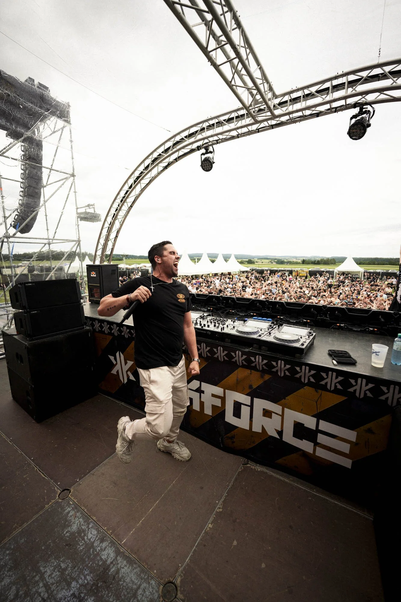 DJ auf einer Bühne bei einem Open-Air-Festival, Publikum im Hintergrund, drinnen und draußen sichtbar, bei Sonnenwetter.