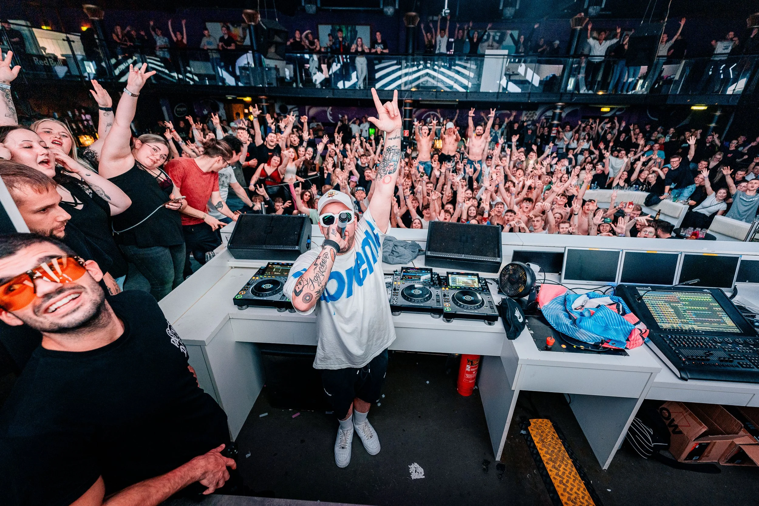 DJ auf einer Bühne in einem Club, umgeben von einem großen Publikum, das elektronische Musik feiert.