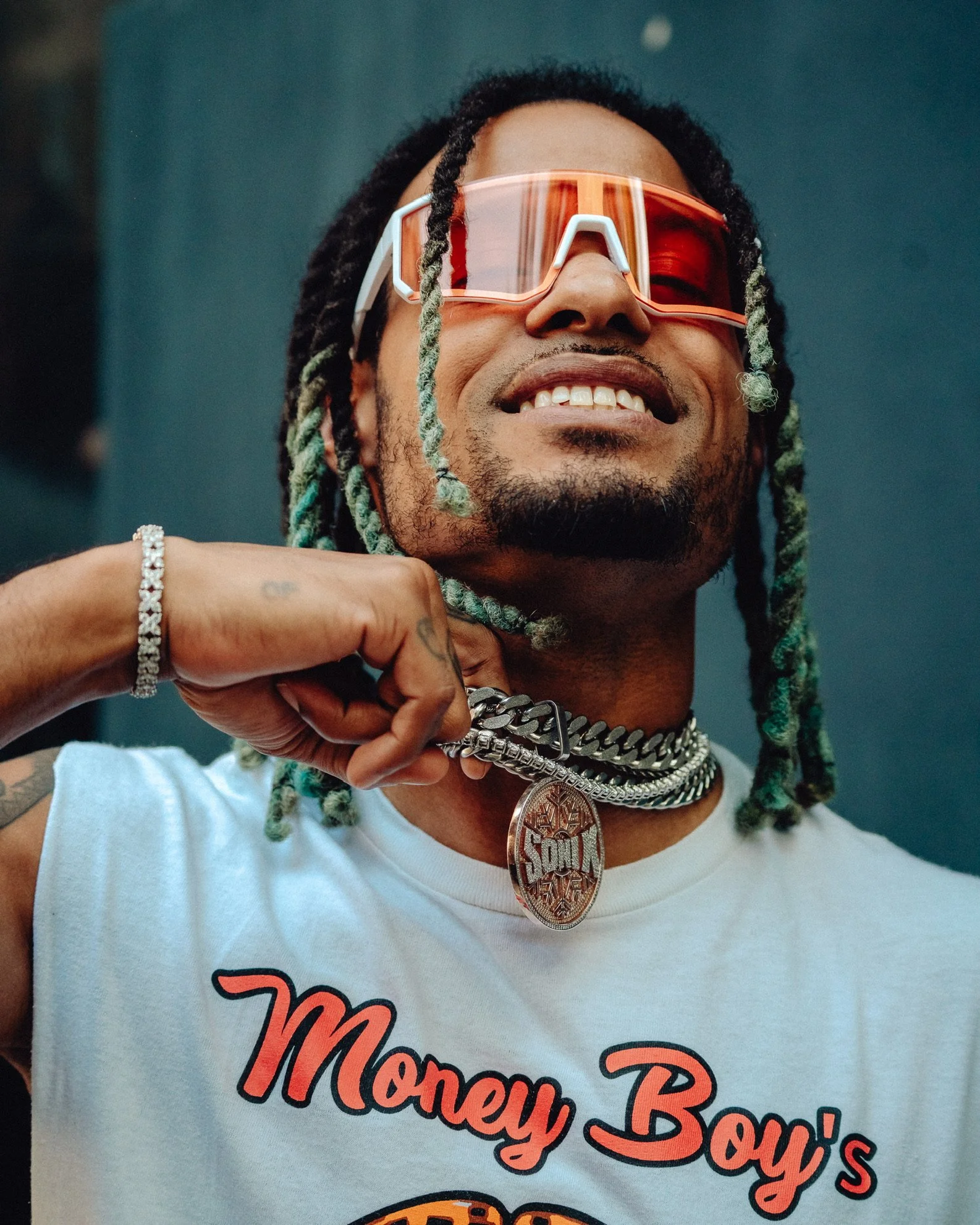 Ein lachender Mann mit Zopffrisur, Sonnenbrille, Schmuck und einem weißen T-Shirt mit der Aufschrift 'Money Boy's'.