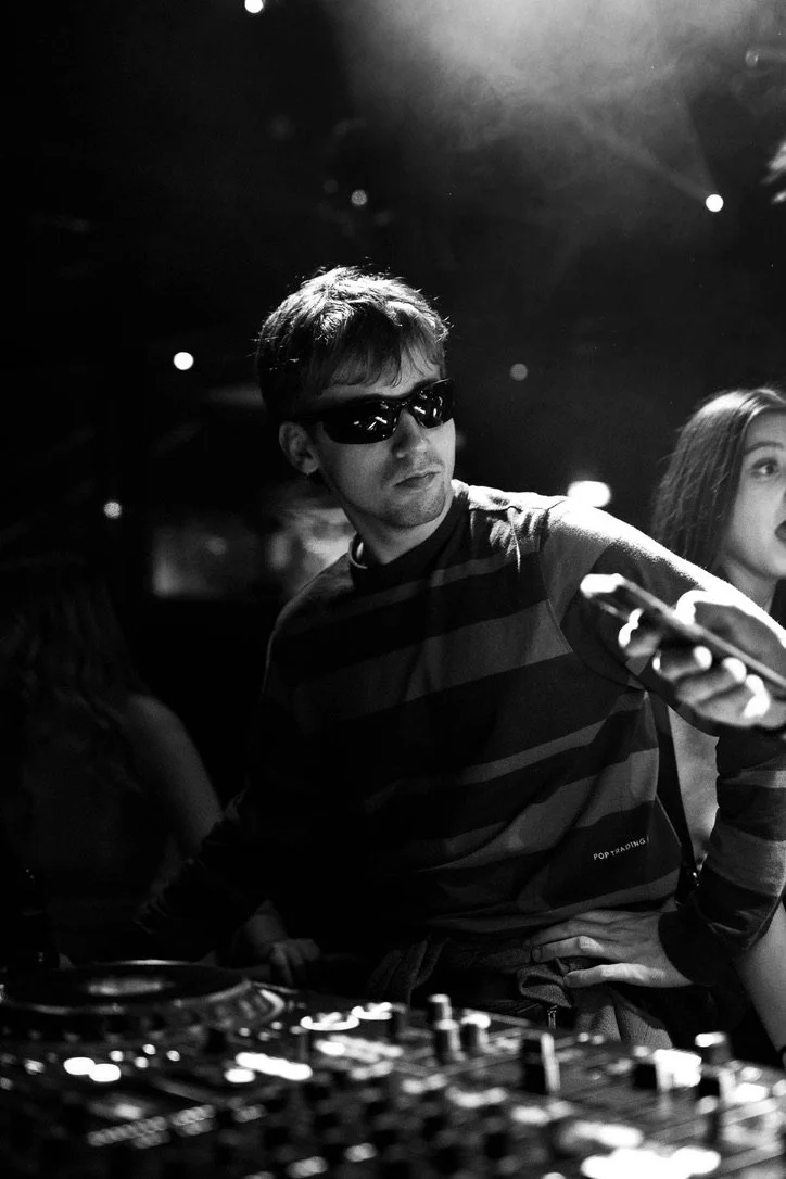 Junge Person mit kurzen Haaren und Sonnenbrille, die in einem dunklen Raum an einem DJ-Setup arbeitet, umgeben von Menschen bei einer Party.