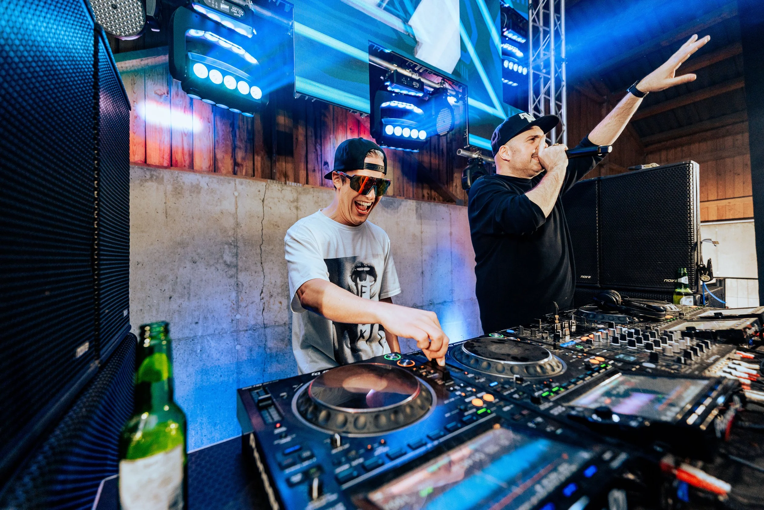 Zwei DJ'S hinter einem Mischpult bei einer Party mit bunten Lichtern und Lautsprechern, eine Person mit Sonnenbrille und Mütze tanzt lachend, die andere Person singt in ein Mikrofon.