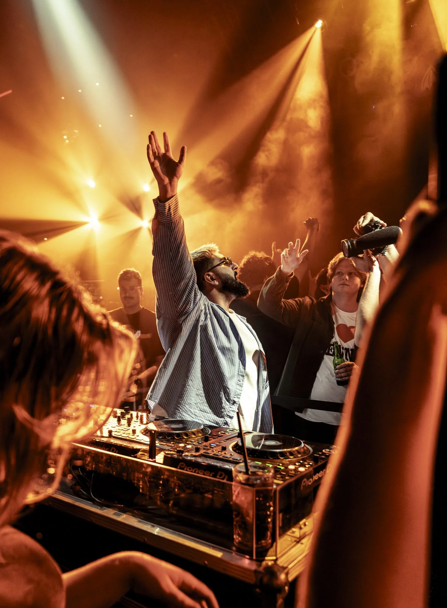 Menschen tanzen auf einer Party vor einer DJ-Konsole, umgeben von orangefarbenen Lichtern und Rauch, mit einem Mann, der die Hände in die Luft wirft.