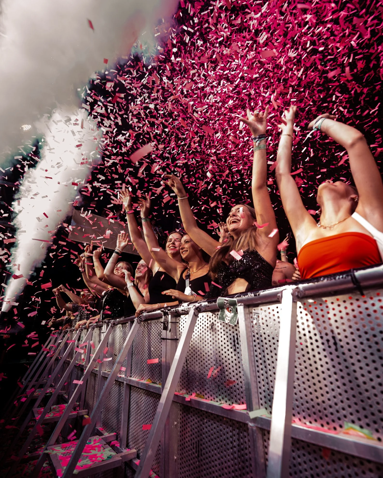 Menschen beim Feiern auf einem Konzert, mit pinkem Konfetti in der Luft, lachende Gesichter und erhobene Arme.