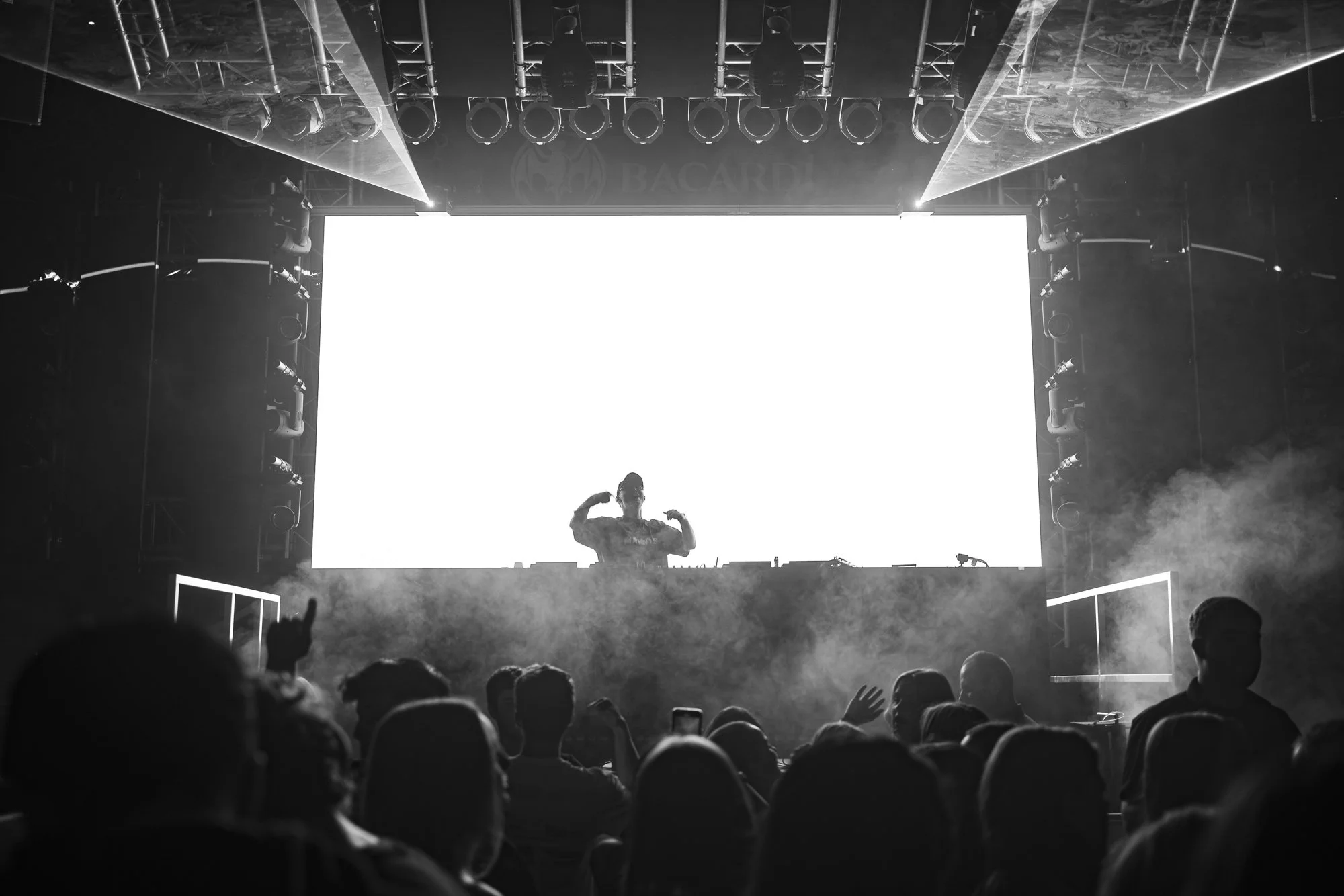 Ein DJ zeigt auf einer Bühne mit einem großen leuchtenden Bildschirm im Hintergrund. Das Publikum schaut zur Bühne, einige erheben die Hände, während es neblig ist.