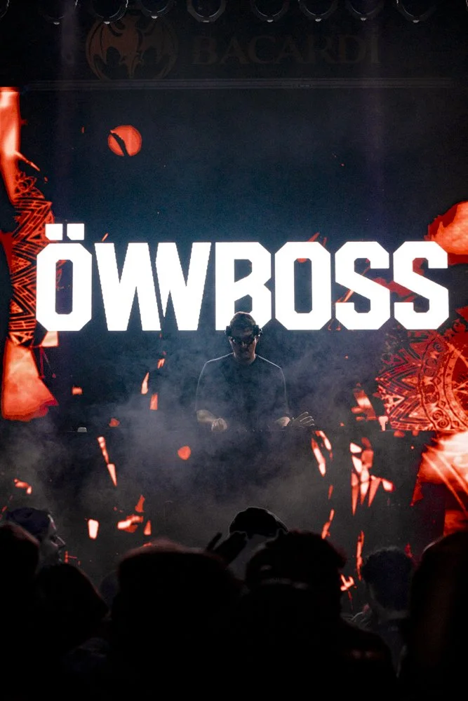 Eine dunkle Bühnenaufnahme mit einem DJ vor einer großen, beleuchteten Anzeige, auf der 'ÖWBOSS' steht, umgeben von roten Lichteffekten und Silhouetten des Publikums.