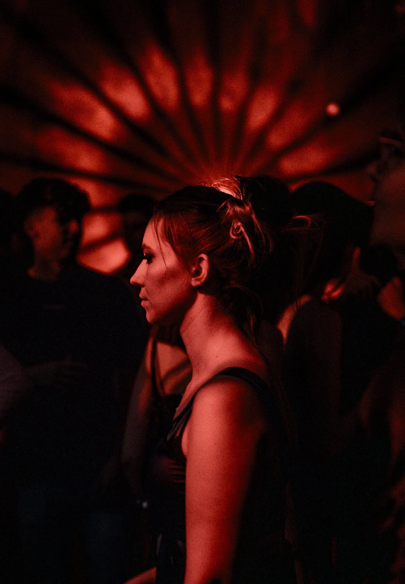 Frau mit roten Haaren in einem schwarzen Kleid in einem dunklen Raum mit rotem Licht, umgeben von anderen Frauen.