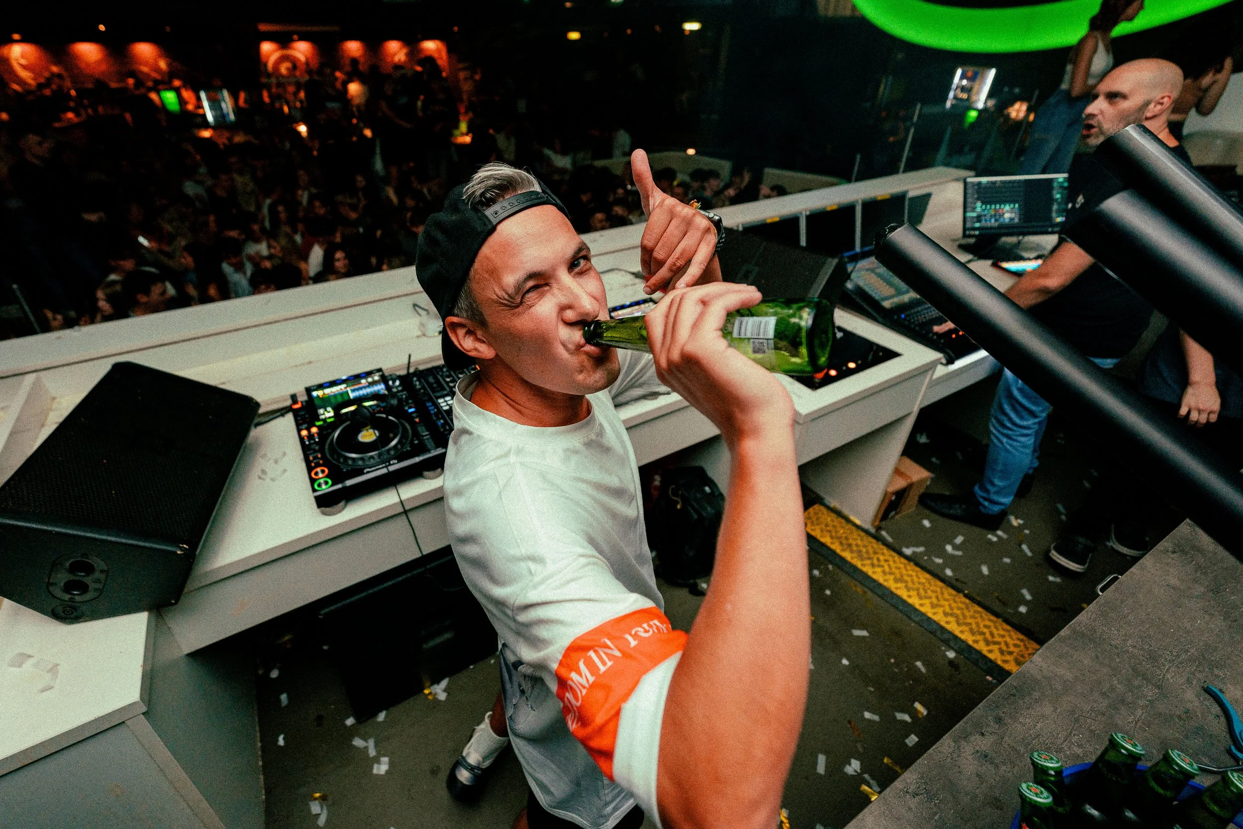 Junger Mann auf einem DJ-Pult, trinkt Bier, winkt in die Kamera, bei einer Party in einem Club, im Hintergrund ein tanzendes Publikum.
