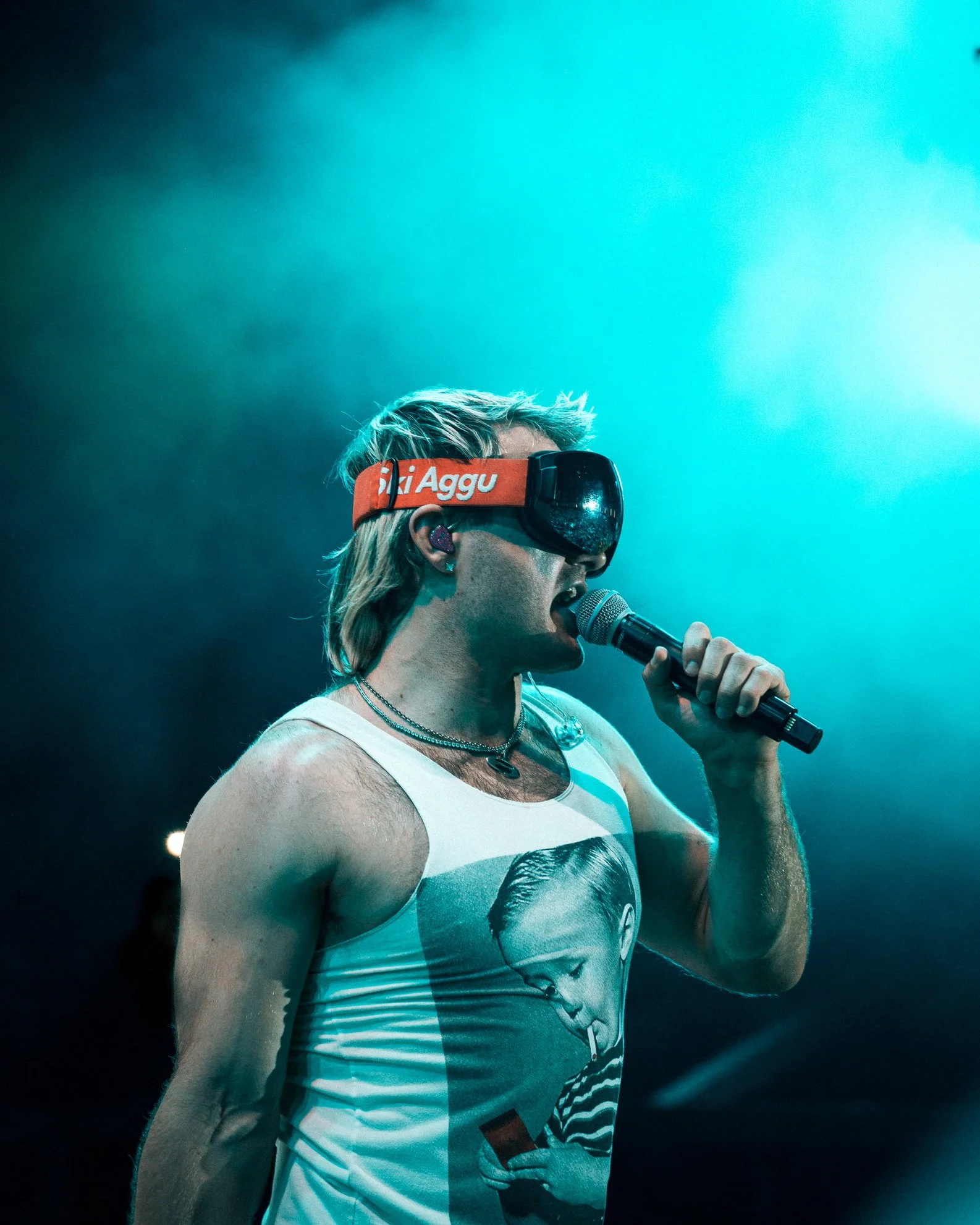 Ein junger Mann, der auf einer Bühne singt, trägt eine Tanktop mit einem abgebildeten Jungen, der auf sein Handy schaut, und eine Virtual-Reality-Brille auf dem Kopf.