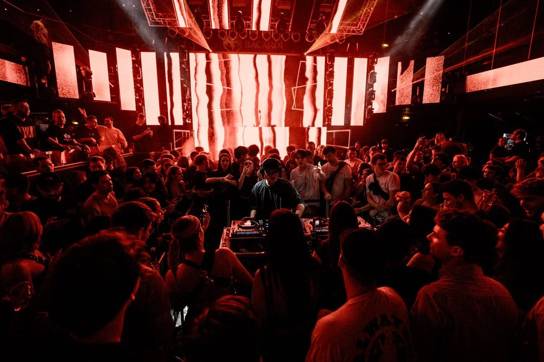 Menschen tanzen und feiern in einem dunklen DJ-Club mit roten Lichtern und LED-Wänden. Ein DJ steht in der Mitte zwischen den Turntables. Die Menge ist begeistert, einige machen Fotos.