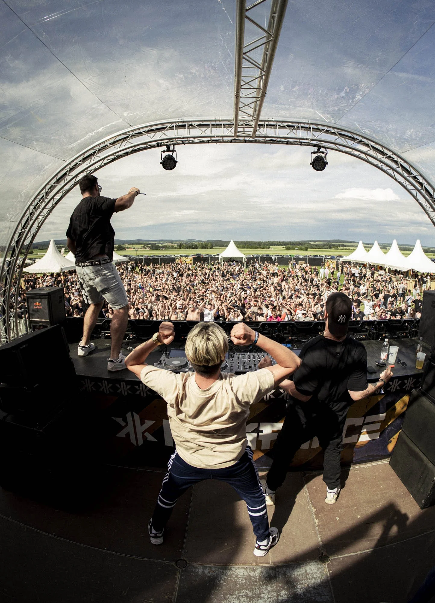 Ein DJ steht auf einer Bühne vor einer großen Menschenmenge bei einem Open-Air-Konzert, mit einem weiten Blick auf die Landschaft im Hintergrund.