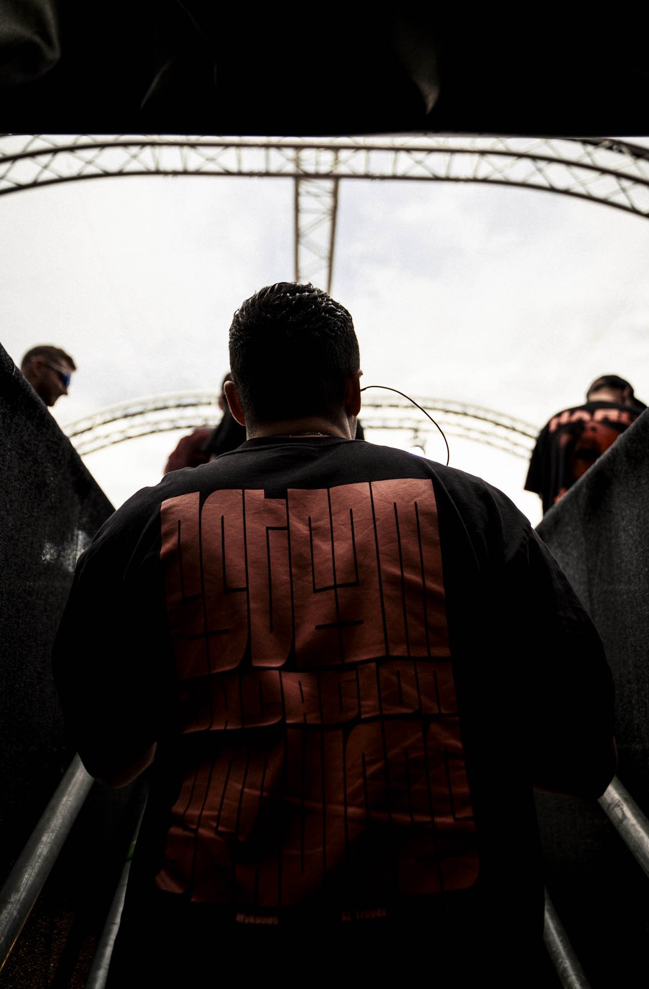 Blick auf einen Mann, der die Treppe zu einem Konzert oder Event hinaufgeht, mit anderen Menschen im Hintergrund, unter einem großen Metallbogen über der Treppe.