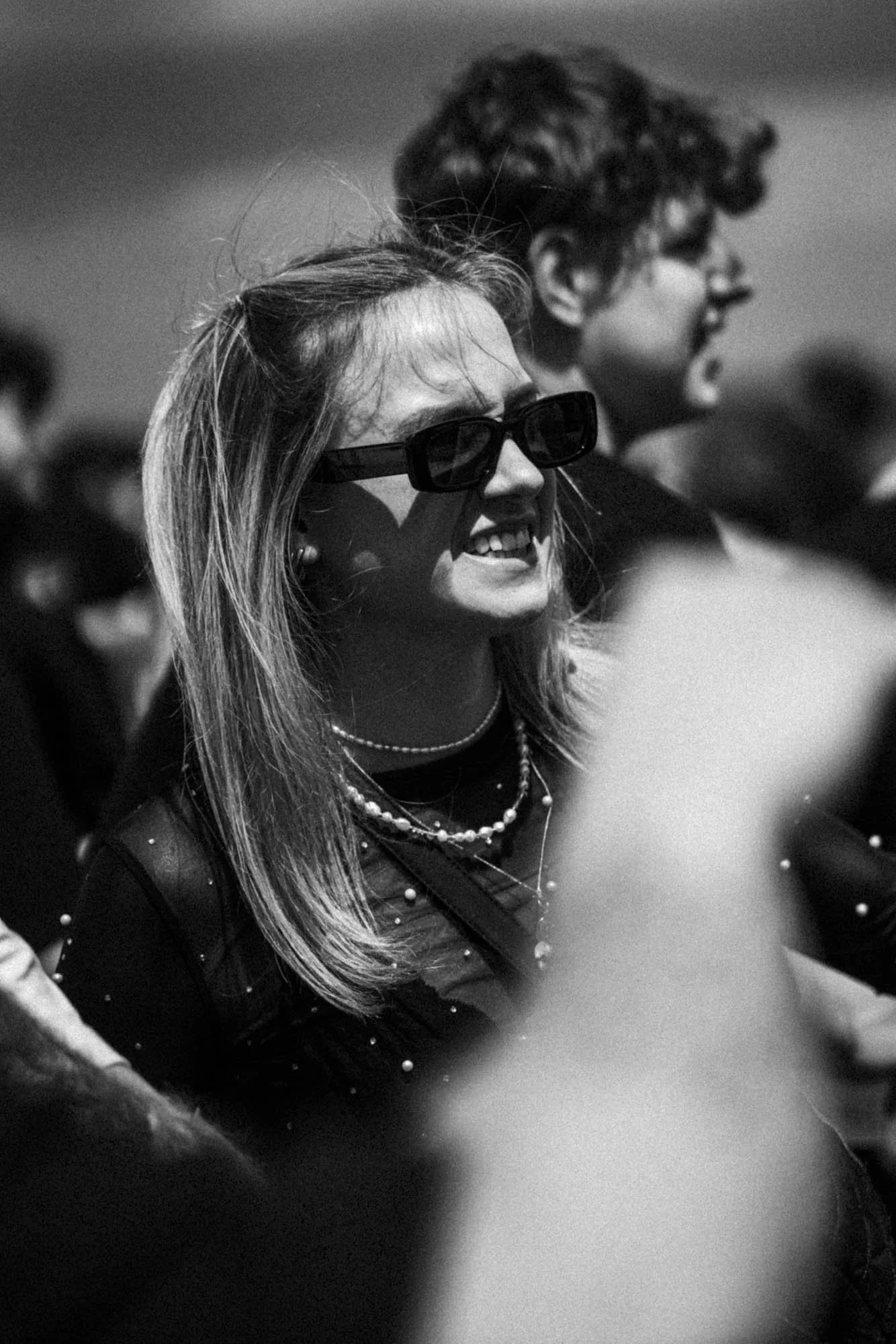 Junge Frau mit langen Haaren und dunklen Sonnenbrillen, lachend während eines Events, mit einem Mann im Hintergrund.