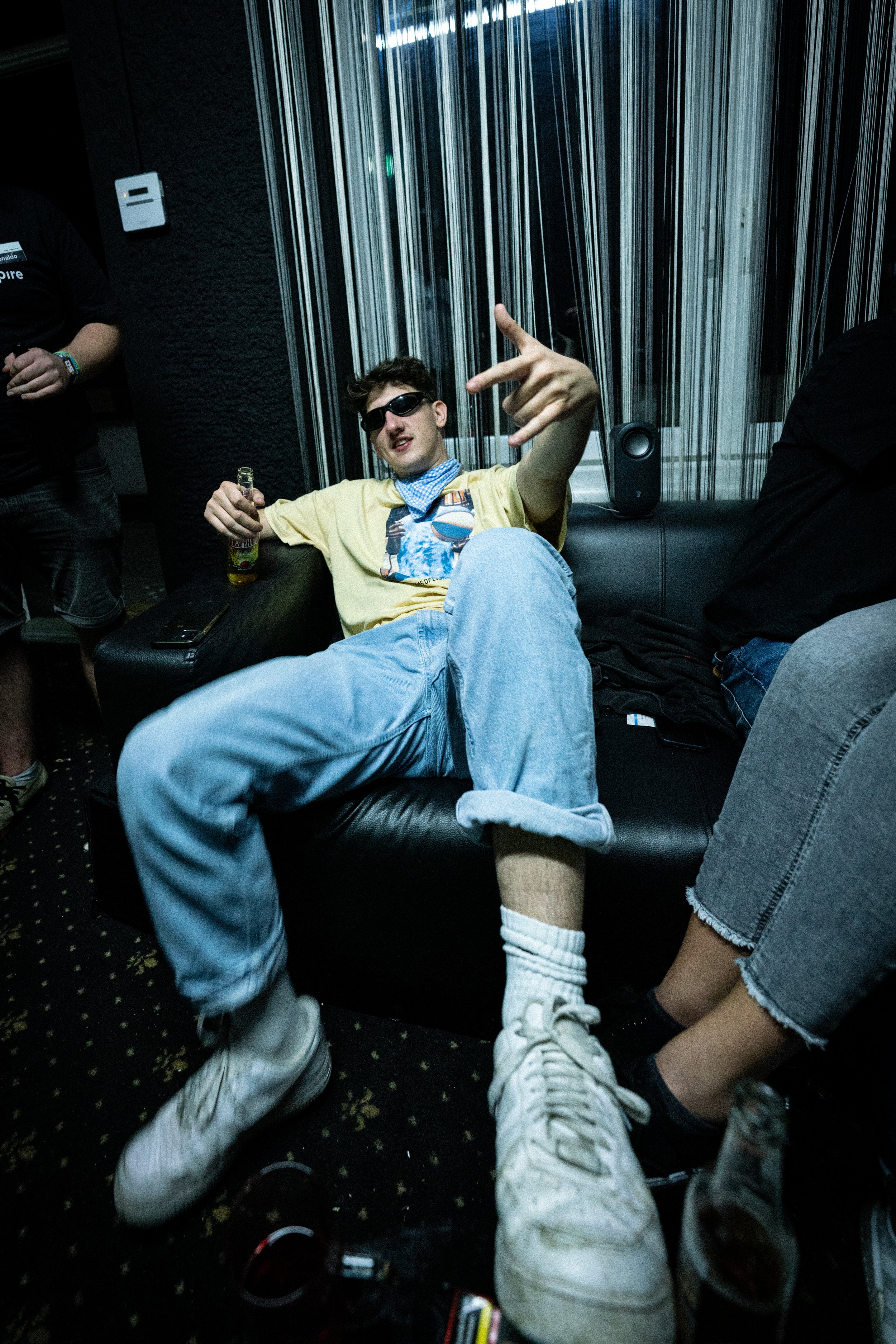 Ein junger Mann sitzt entspannt auf einem schwarzen Sofa, trägt dunkle Sonnenbrillen, ein gelbes T-Shirt mit einem bunten Druck, blaue Jeans, weiße Turnschuhe, weiße Socken und hält eine Flasche. Er macht eine Handgeste und lächelt, umgeben von ander
