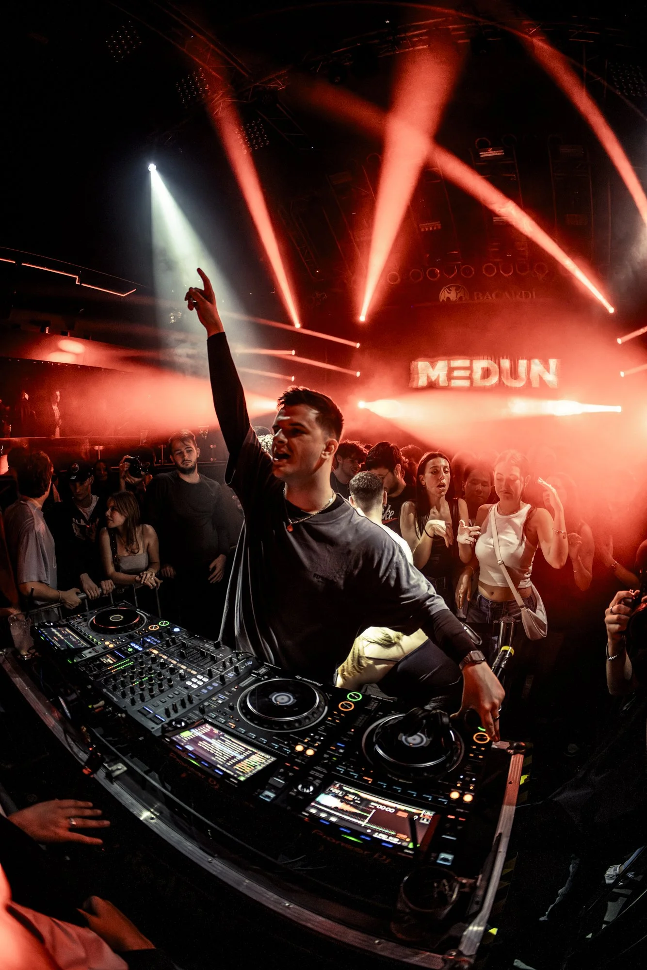 Ein DJ steht an einer DJ-Konsole in einer Club-Atmosphäre mit roten Lichtern und einer Tanzfläche voller Menschen, die feiern.