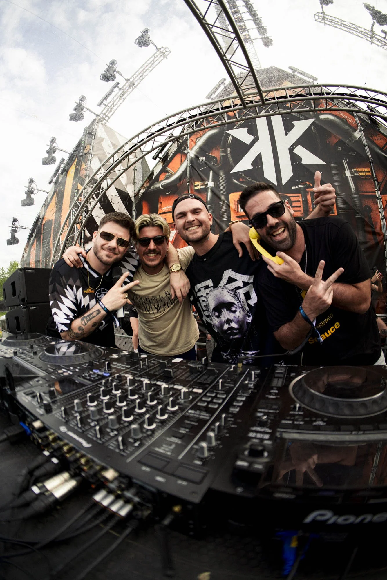 Vier Männer auf einer DJ-Stage bei einem Musikfestival, lachend und mit Handzeichen, im Hintergrund ein Bühnenaufbau mit Lichtelementen.