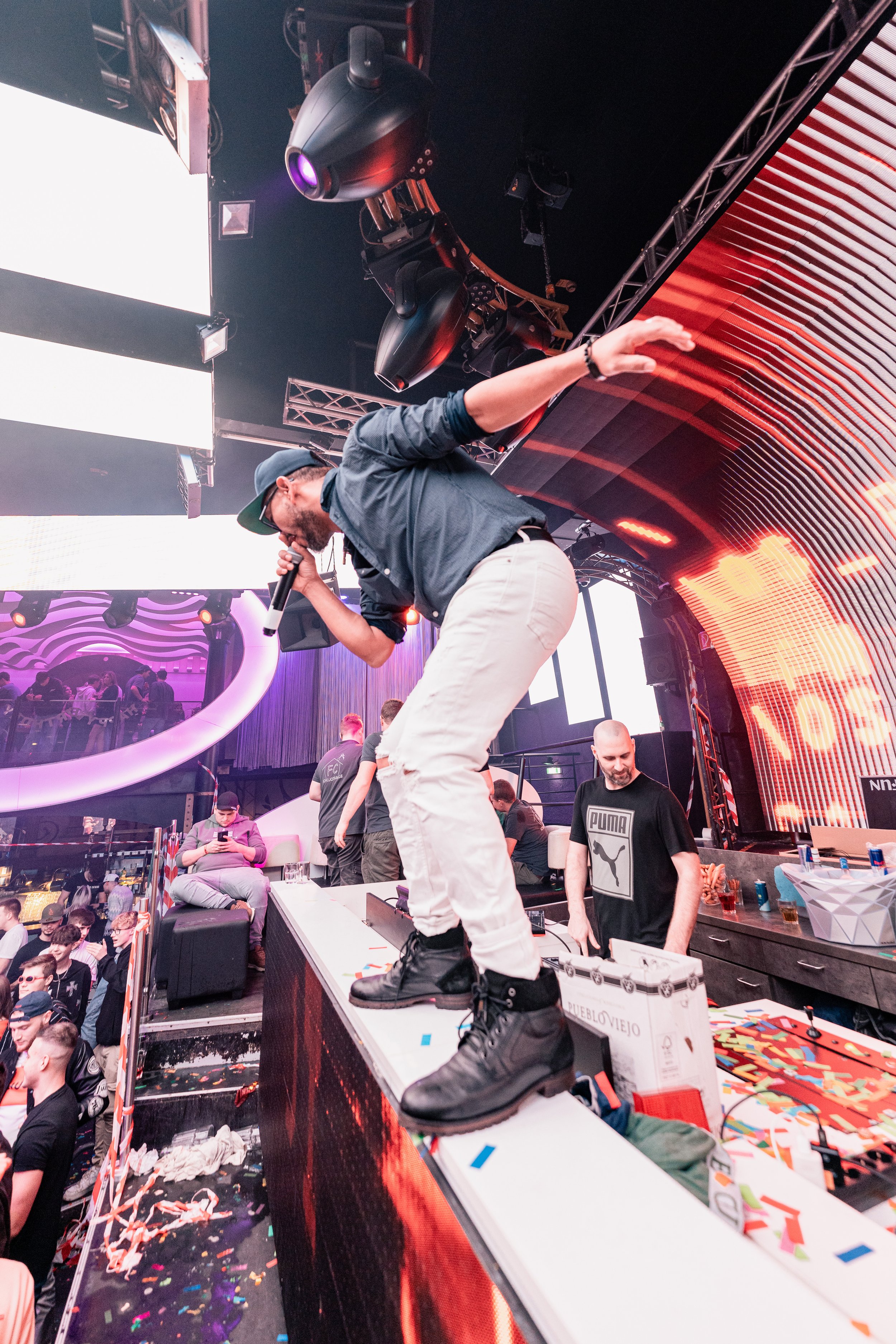 Ein Mann tanzt auf einer Bühne bei einer Party, umgeben von anderen Menschen und bunten Lichtern.