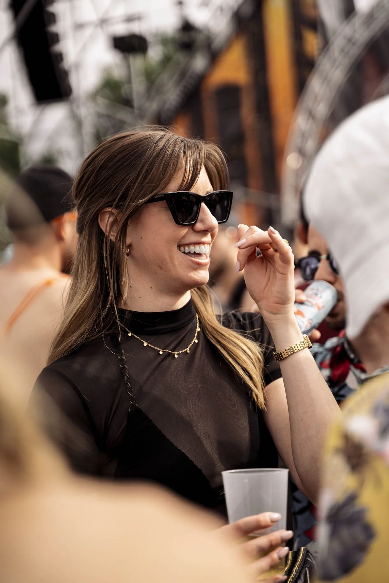 Eine lachende Frau mit Sonnenbrille und goldener Uhr bei einem Event im Freien.