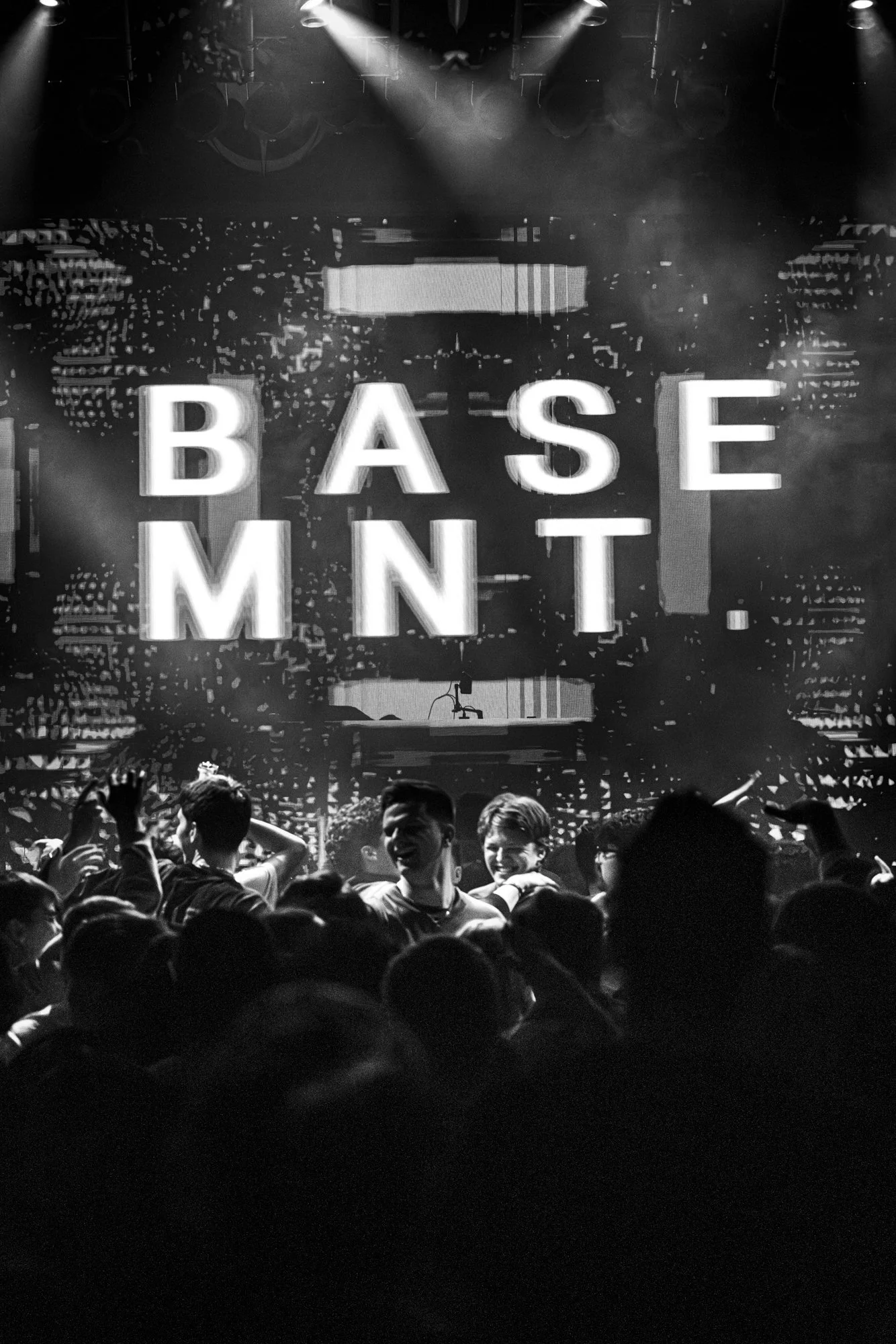 Menschen tanzen und feiern auf einer schwarzen und weißen Konzertbühne mit großen leuchtenden Buchstaben, die 'BASE MNT' zeigen.