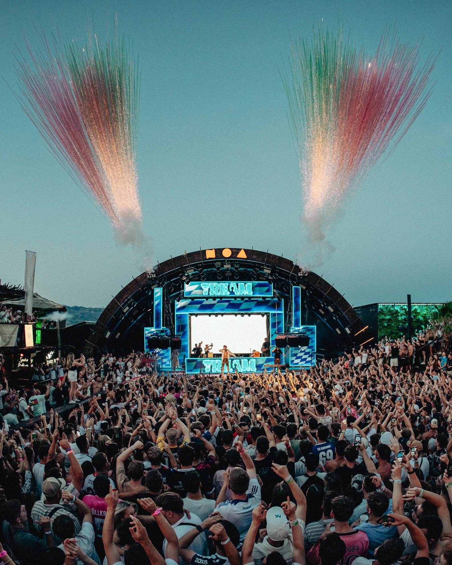 Menschenmenge beim Konzert auf einer Open-Air-Bühne mit Farbenfeuerwerk am Himmel.