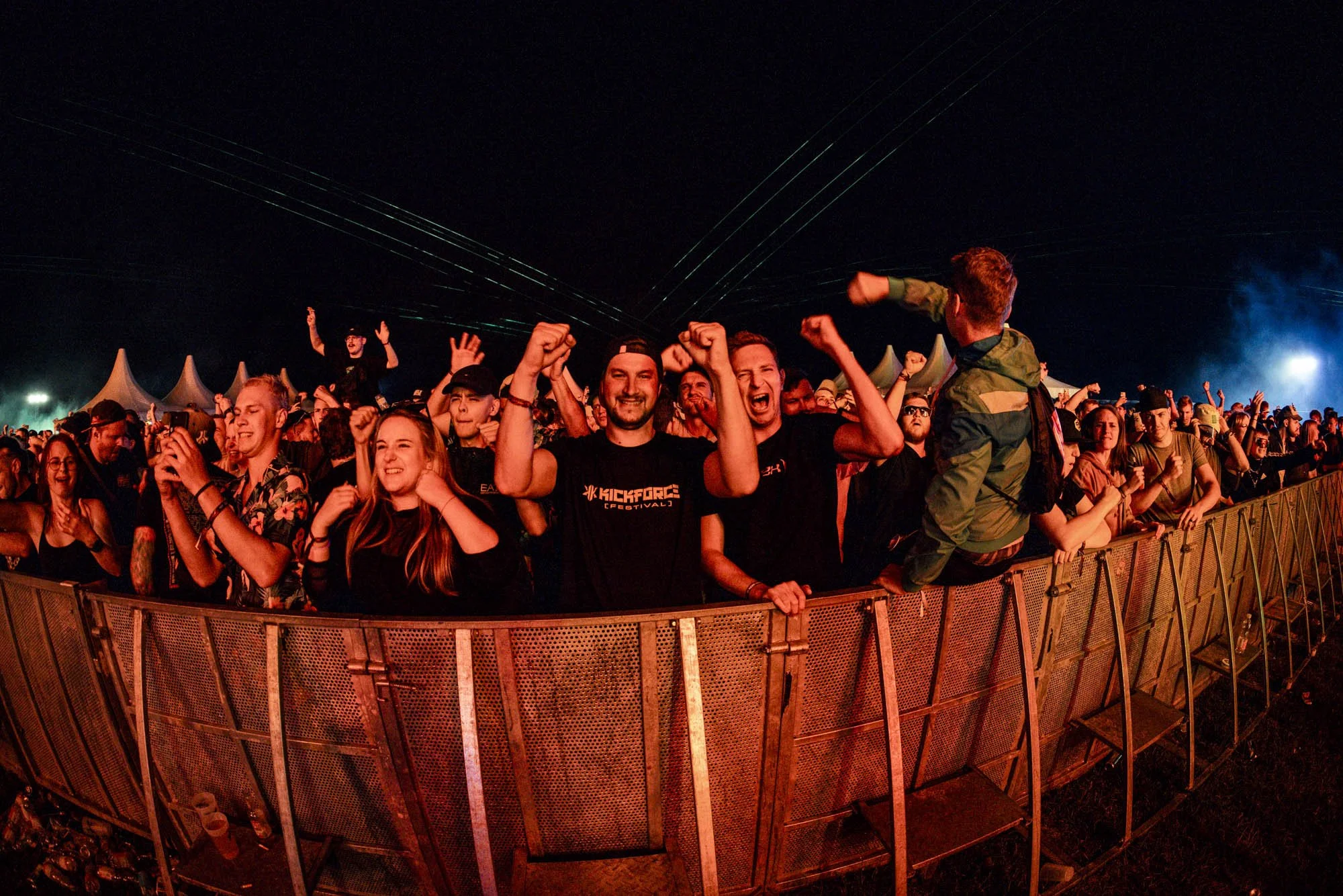 Menschen beim Musikfestival, laut feiernd, mit Fanfaren und Zelten im Hintergrund, nachts mit Lichter und Bands.