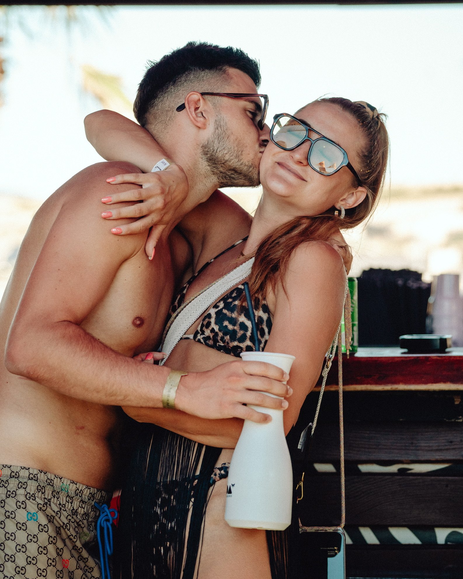 Ein junger Mann und eine Frau bei einem Strandbesuch, umarmen sich, der Mann küsst die Frau auf die Wange, die Frau trägt eine Sonnenbrille und ein Tiermuster-Bikini, sie hält eine weiße Flasche, im Hintergrund sind Sonnenschirme und Strandzubehör si