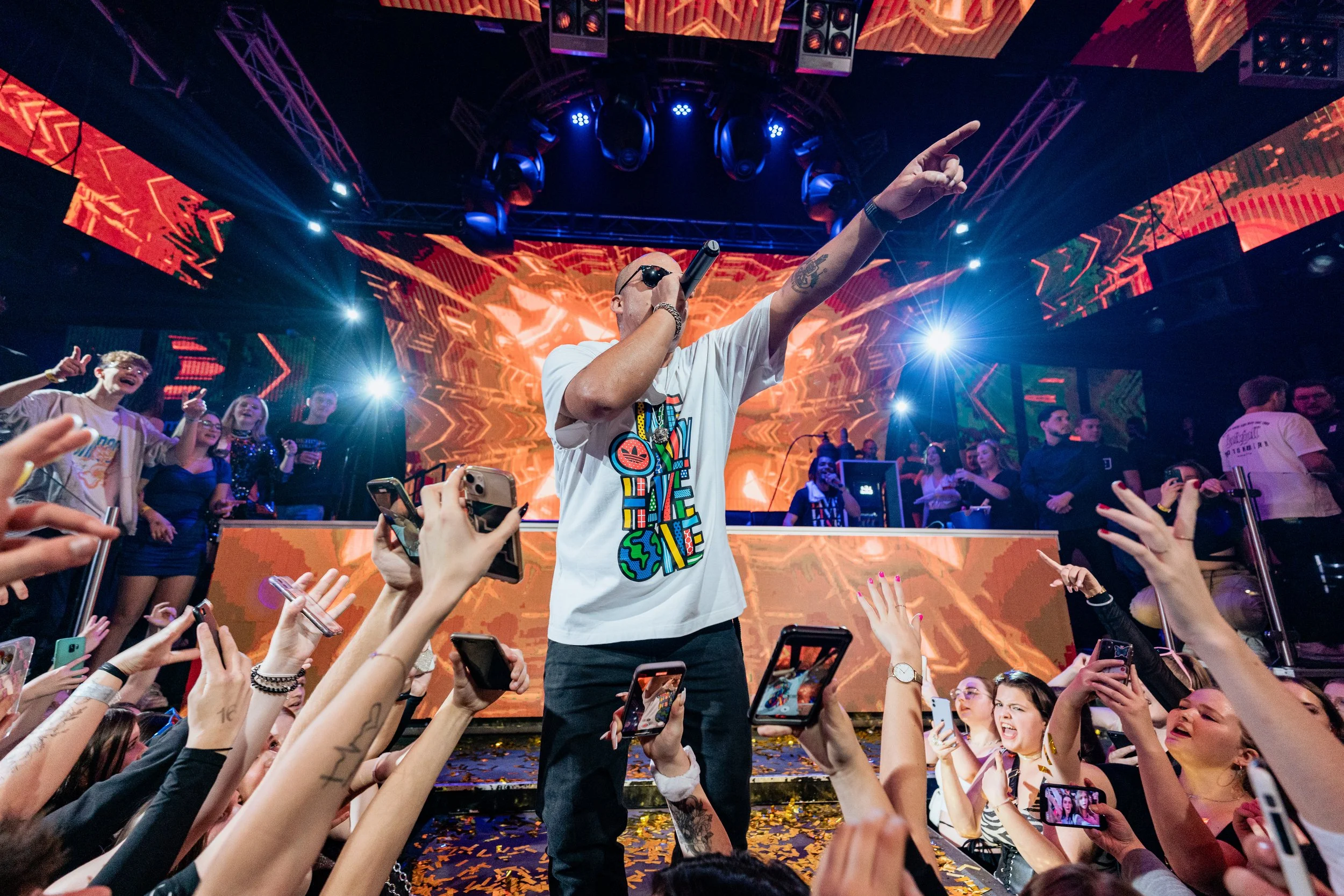 Ein männlicher Performer auf einer Bühne, umgeben von einem Publikum mit Smartphones, beim Live-Konzert oder DJ-Event, mit bunten Lichtern und LED-Bildschirm im Hintergrund.