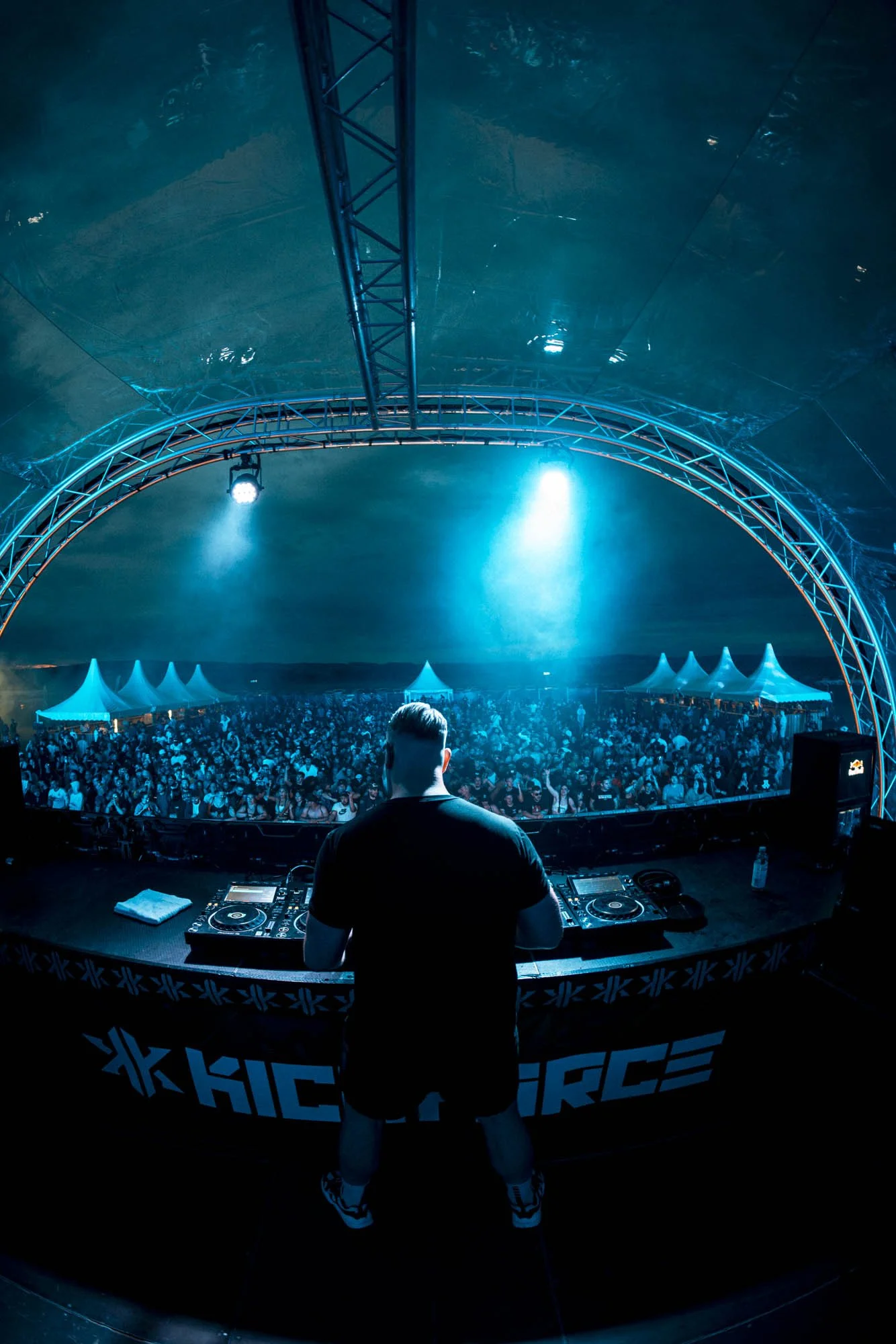 Ein DJ spielt auf einer Bühne vor einer großen Menschenmenge bei einem Musikfestival bei Nacht, mit blauen Lichter und weißen Zelten im Hintergrund.