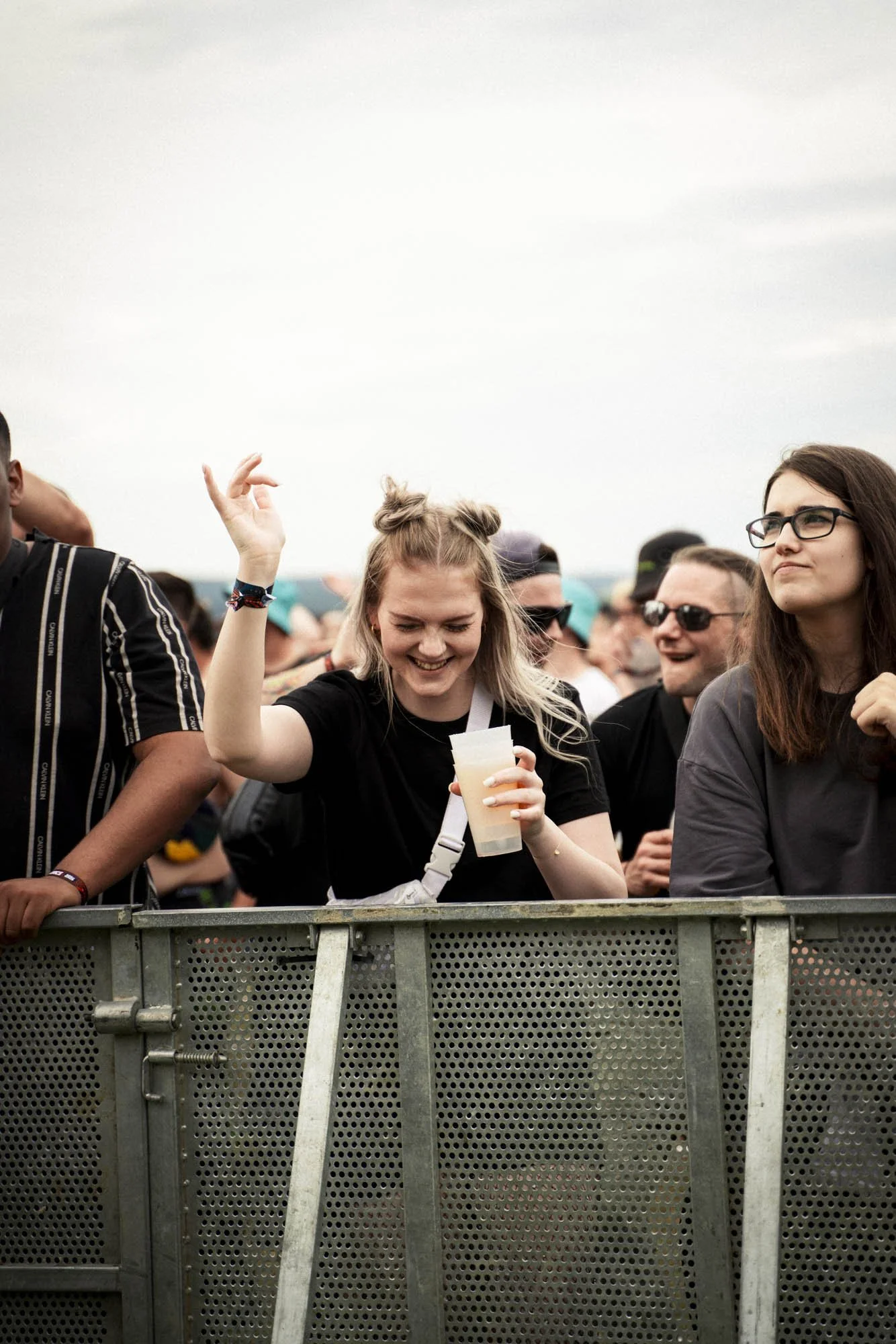 Menschen stehen an einem Konzertzaun, die junge Frau in der Mitte lächelt und hebt die Hand beim Tanzen, hält ein Getränk in der anderen Hand; weitere Menschen im Hintergrund bei einem Musikfestival.