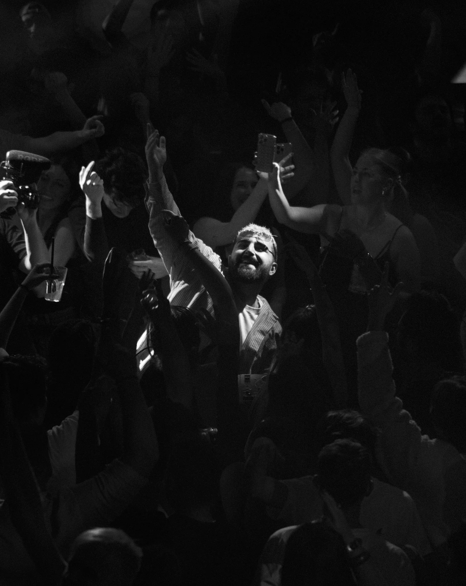 Menschenmenge bei Konzert, eine Person im Zentrum, die in die Luft zeigt und eine andere Person, die ein Handy hält, alle in dunkler Beleuchtung.