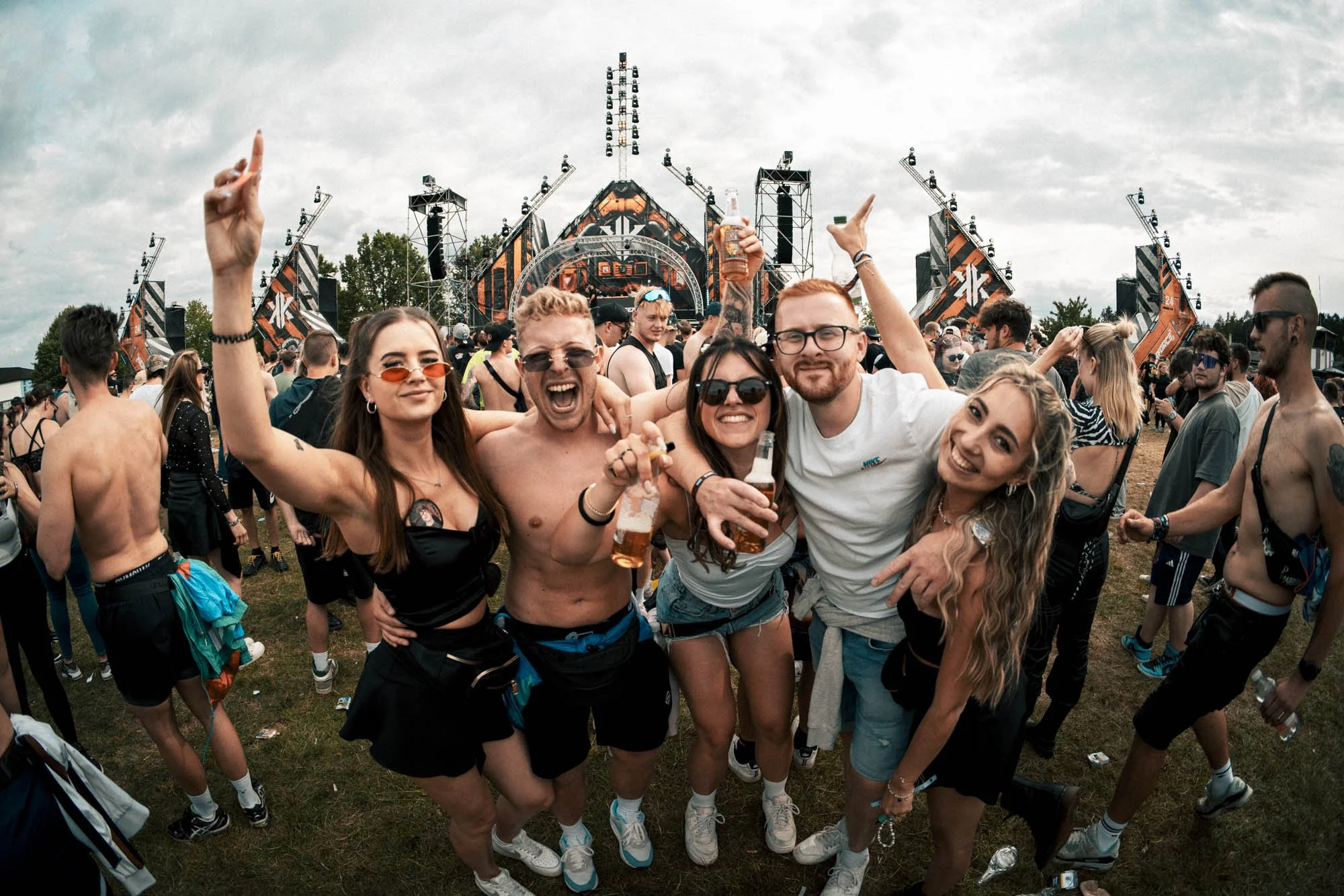 Gruppe von fünf jungen Menschen, die auf einem Festival fröhlich in die Kamera lächeln, einige mit Bierflaschen in den Händen, im Hintergrund eine große Bühne und viele weitere Festivalbesucher.