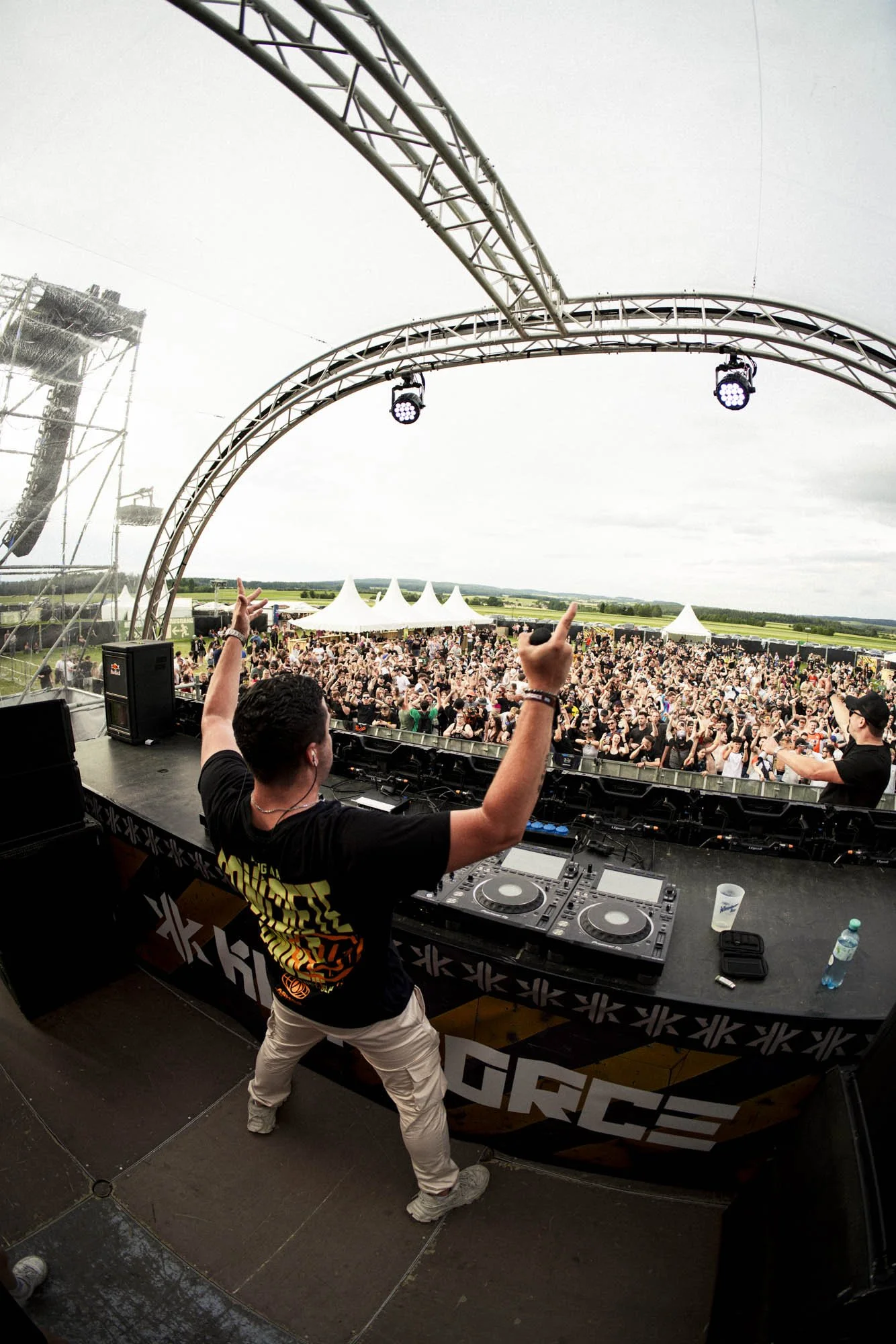 DJ auf einer Bühne bei einem Open-Air-Festival, mit einem großen Publikum vor der Bühne, einige Zuschauer heben die Hände, im Hintergrund Zelte und grüne Felder unter einem bewölkten Himmel.