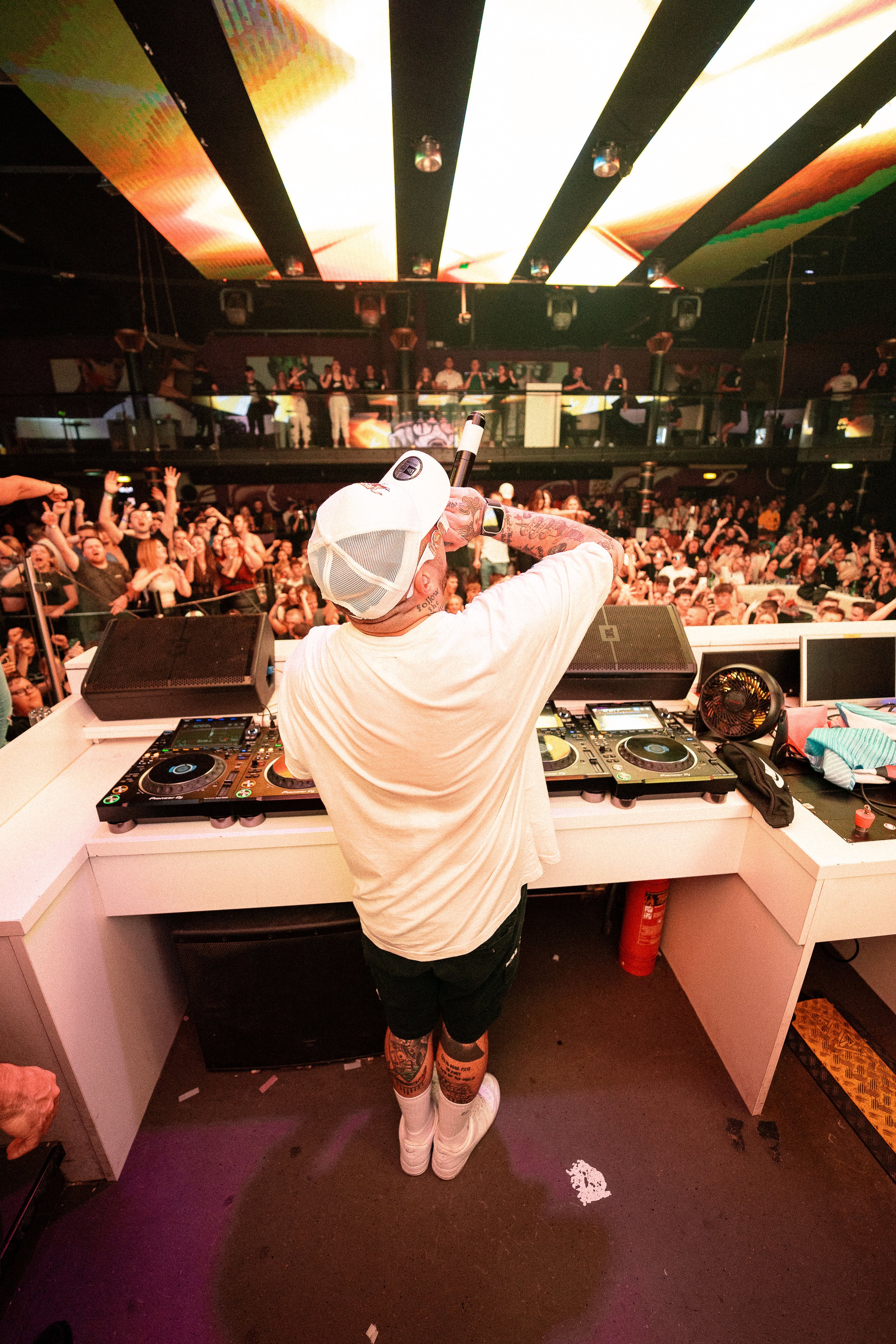 Ein DJ tanzt an seinem DJ-Pult in einer vollen Diskothek mit vielen tanzenden Gästen unter einer großen, bunten Lichtinstallation an der Decke.