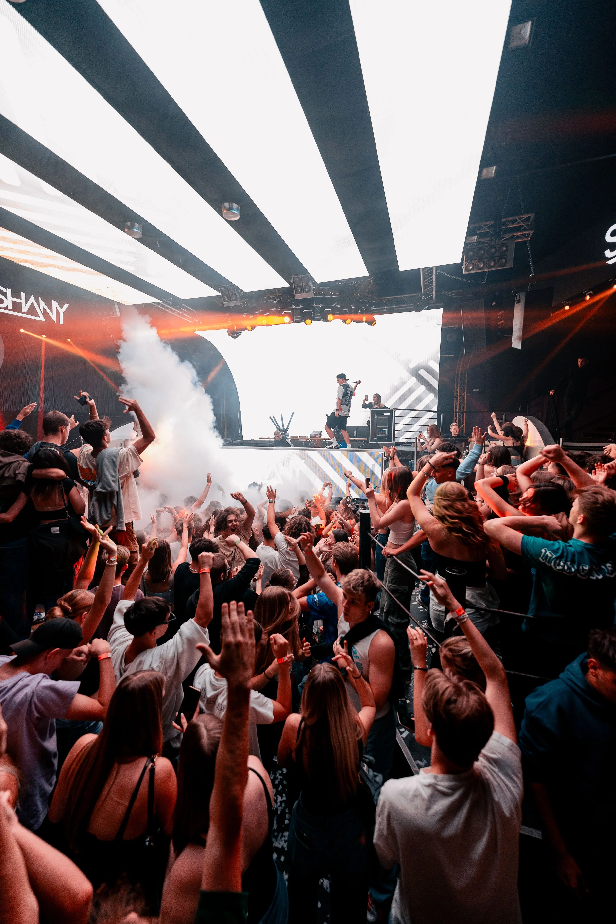 Eine Menschenmenge tanzt und feiert bei einem Konzert oder Festival in einer dunklen Halle mit bunten Lichtern und Nebel. Auf der Bühne sind Musiker oder DJs zu sehen, die aufführen.