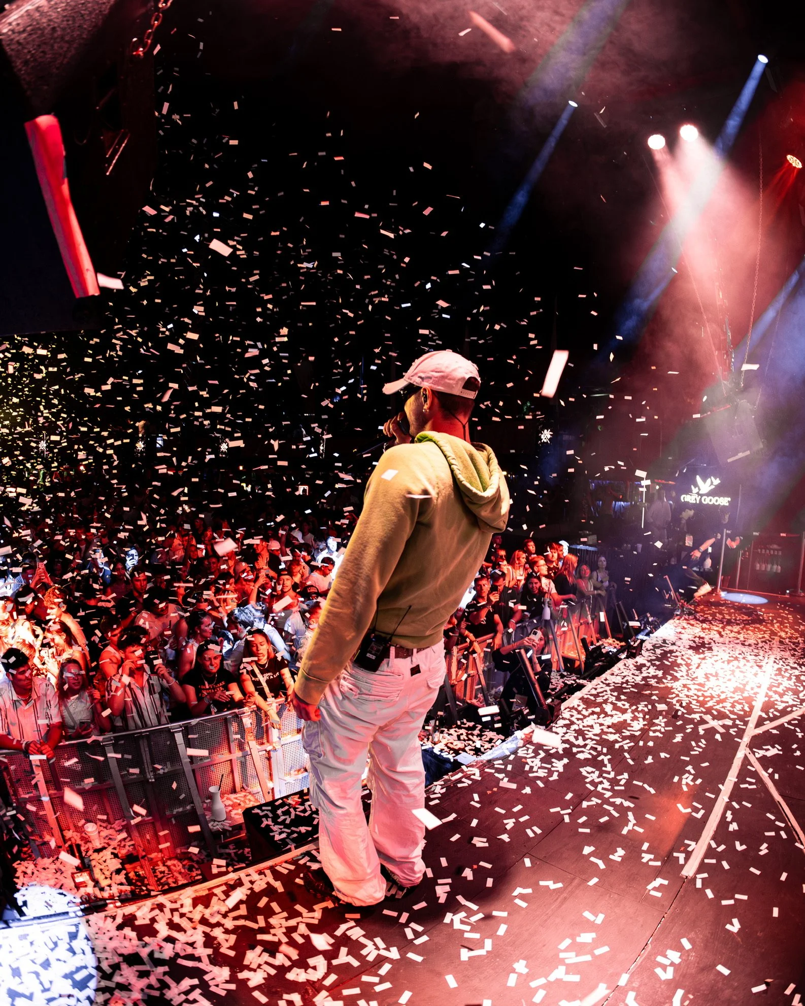Ein Sänger steht auf einer Bühne, während Konfetti auf das Publikum fällt, bei einem Live-Konzert in einer dunklen, beleuchteten Club- oder Theaterszene mit rotem und blauem Licht.