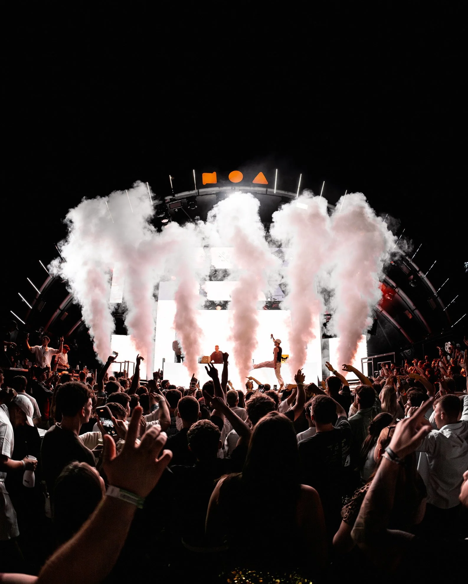 Auf einer Musikbühne bei einem Konzert werden Rauch- und Pyrotechnik-Effekte eingesetzt, mit einem Publikum im Vordergrund, das tanzt und fotografiert.