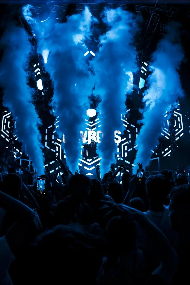 DJ auf einer Bühne bei einem Musikfestival, umgeben von Rauch und blauen Lichteffekten, Menschen im Publikum nehmen Fotos auf.