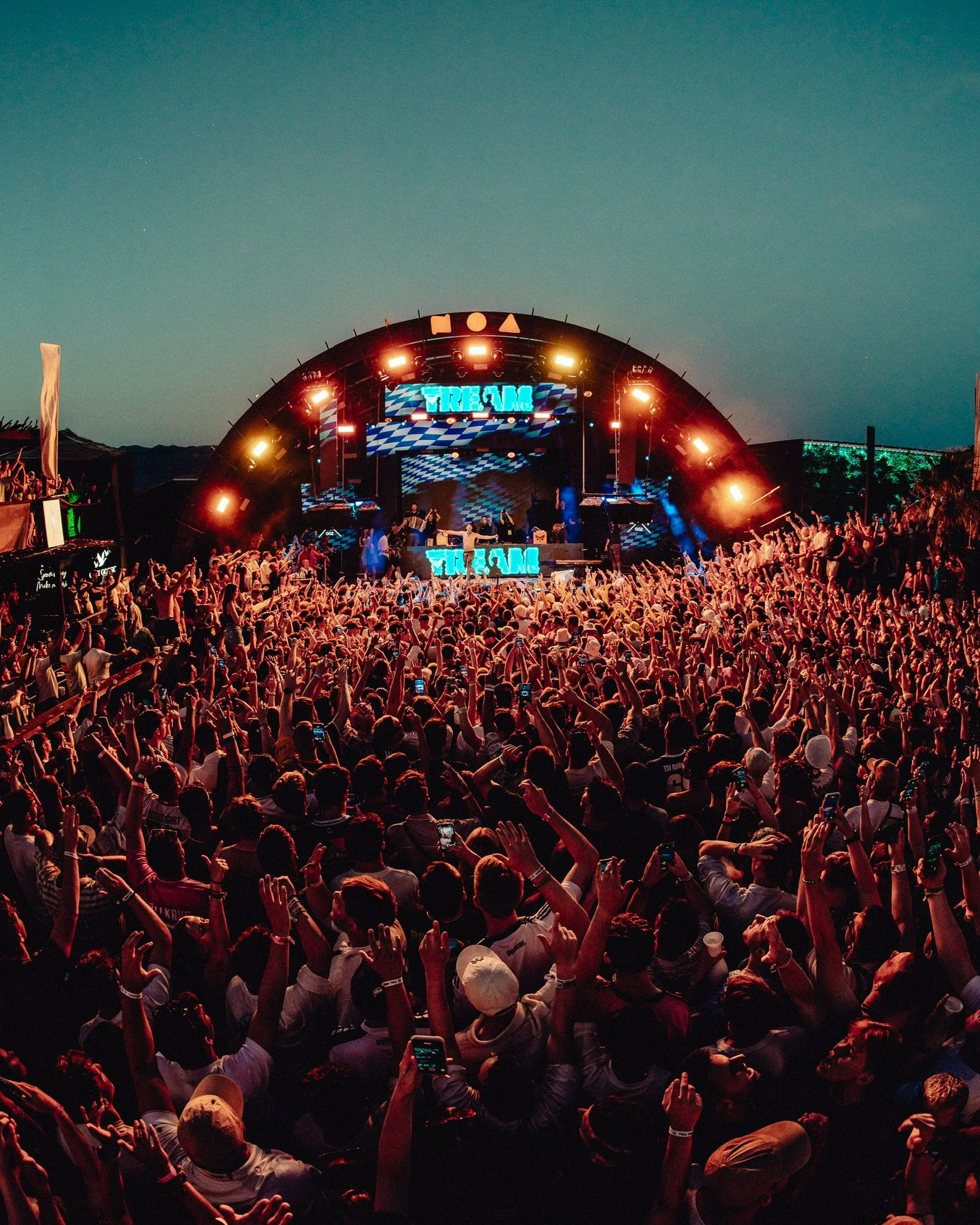 Menschenmengen vor einer großen Bühne bei einem Open-Air-Konzert mit Beleuchtung und einem dunklen Himmel im Hintergrund.