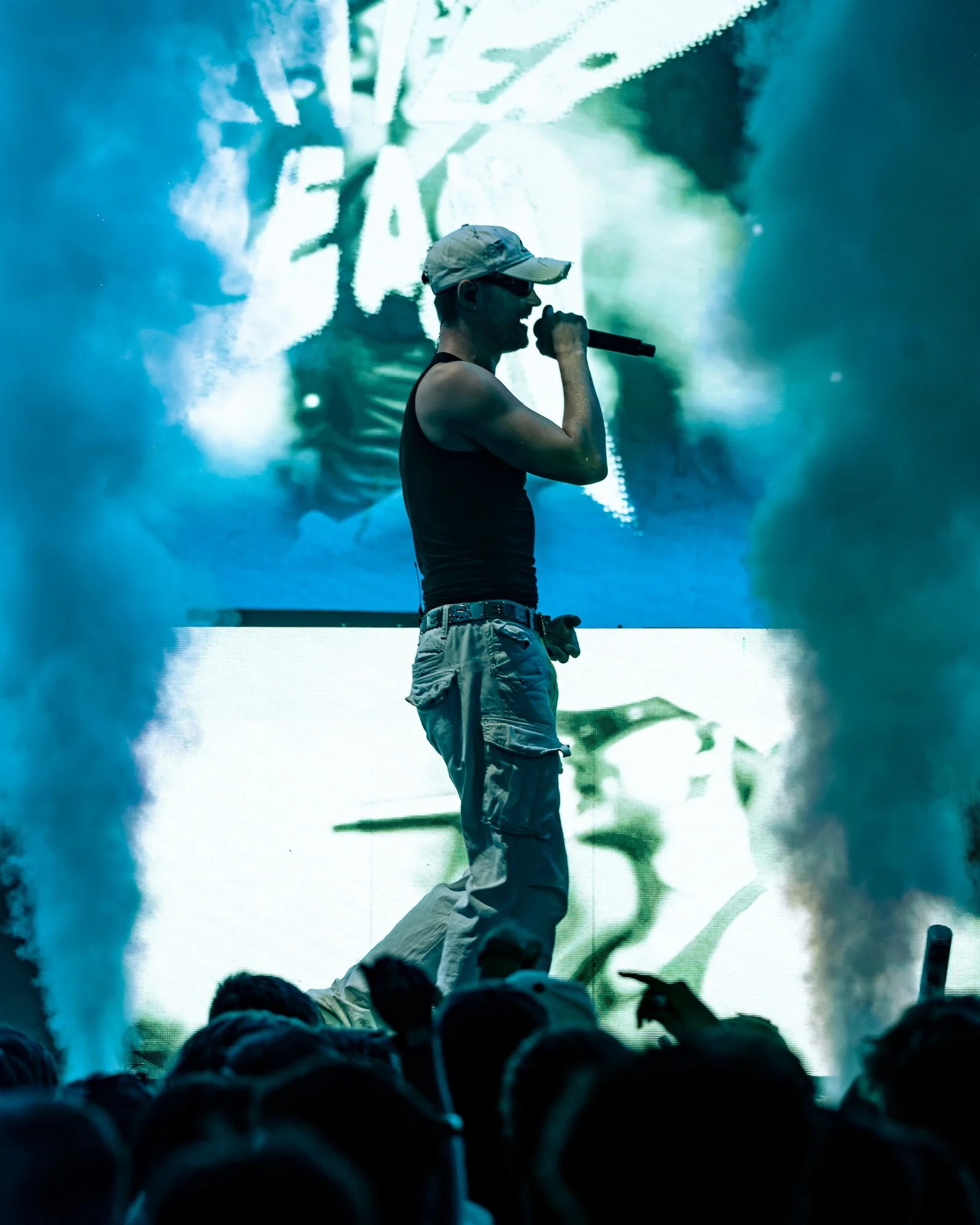 Ein männlicher Musiker in dunklen Kleidern mit Sonnenbrille und Basecap singt in ein Mikrofon auf einer Bühne, während ein großes Bildschirm im Hintergrund mit einer abstrakten Grafik oder Bild zu sehen ist. Das Publikum im Vordergrund ist sichtbar.