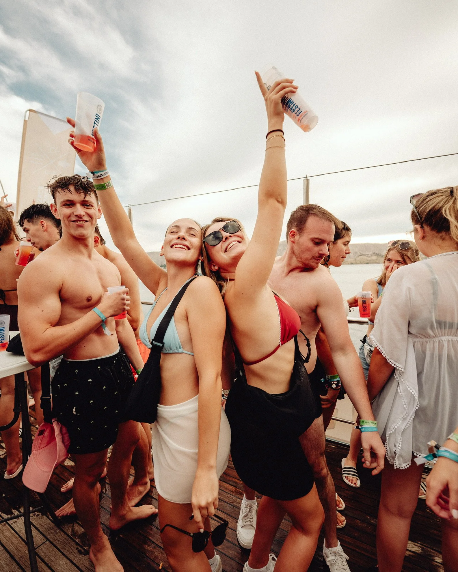 Junge Menschen feiern auf einer Party im Freien, einige tragen Bikinis und Shorts, heben Getränke in die Luft, lachen und haben Spaß.