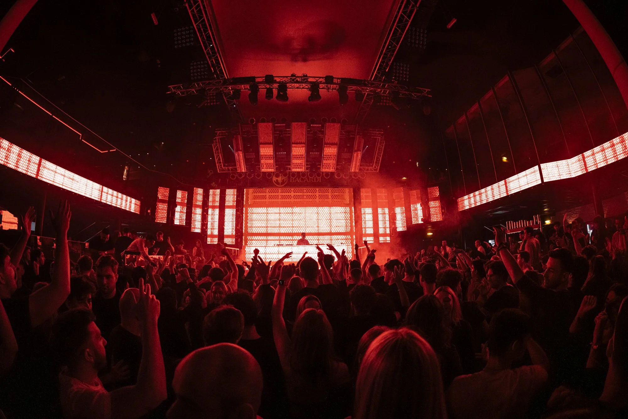 Menschen tanzen bei einem Konzert oder einer Nachtclub-Party mit roter Beleuchtung und großem LED-Bildschirm im Hintergrund.