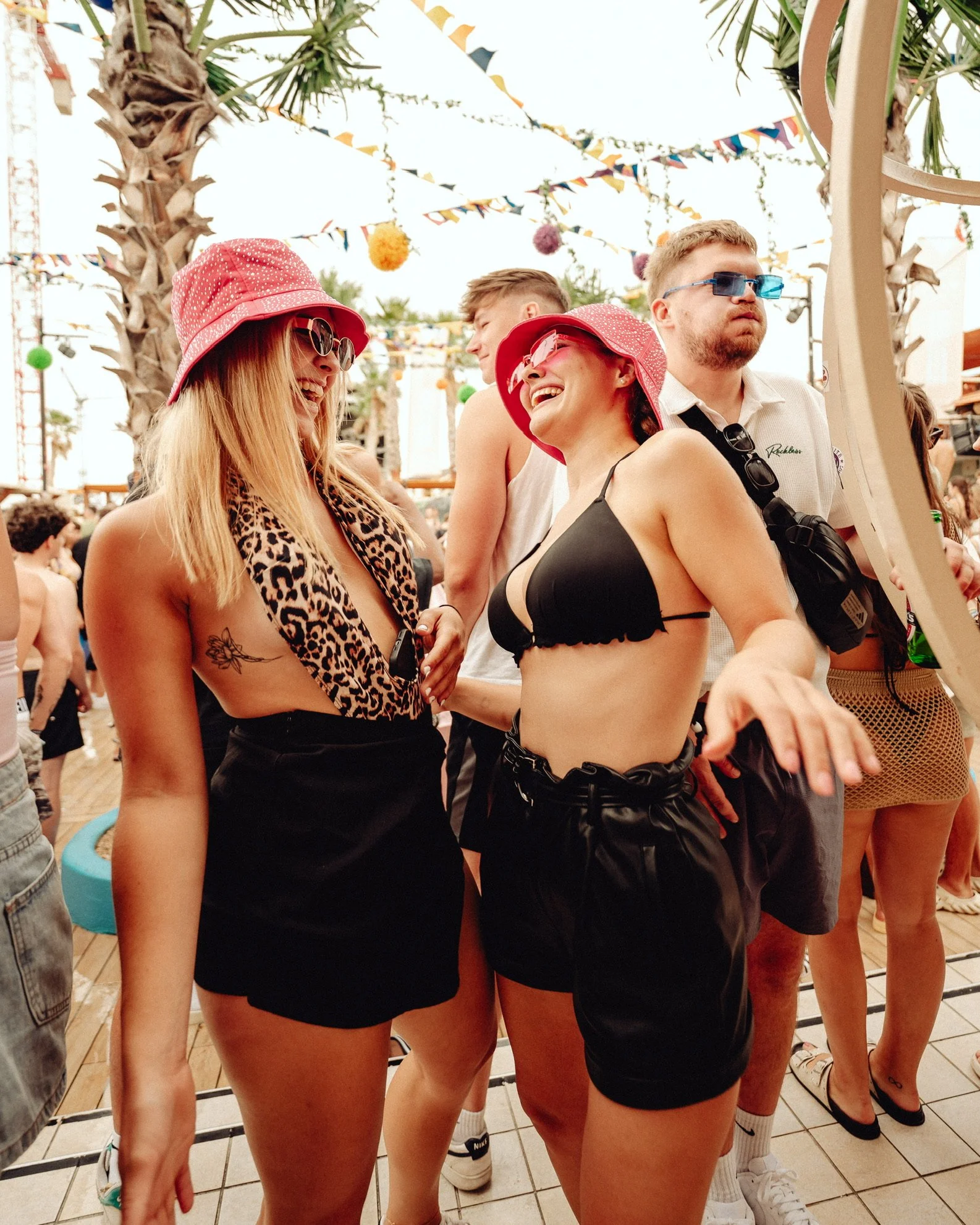 Zwei lachende Frauen beim Festival, eine trägt einen Leoparden-Print-Schal und einen pinken Hut, die andere einen pinken Hut und schwarze Shorts, umgeben von weiteren Festivalbesuchern unter bunter Dekoration und Palmen.