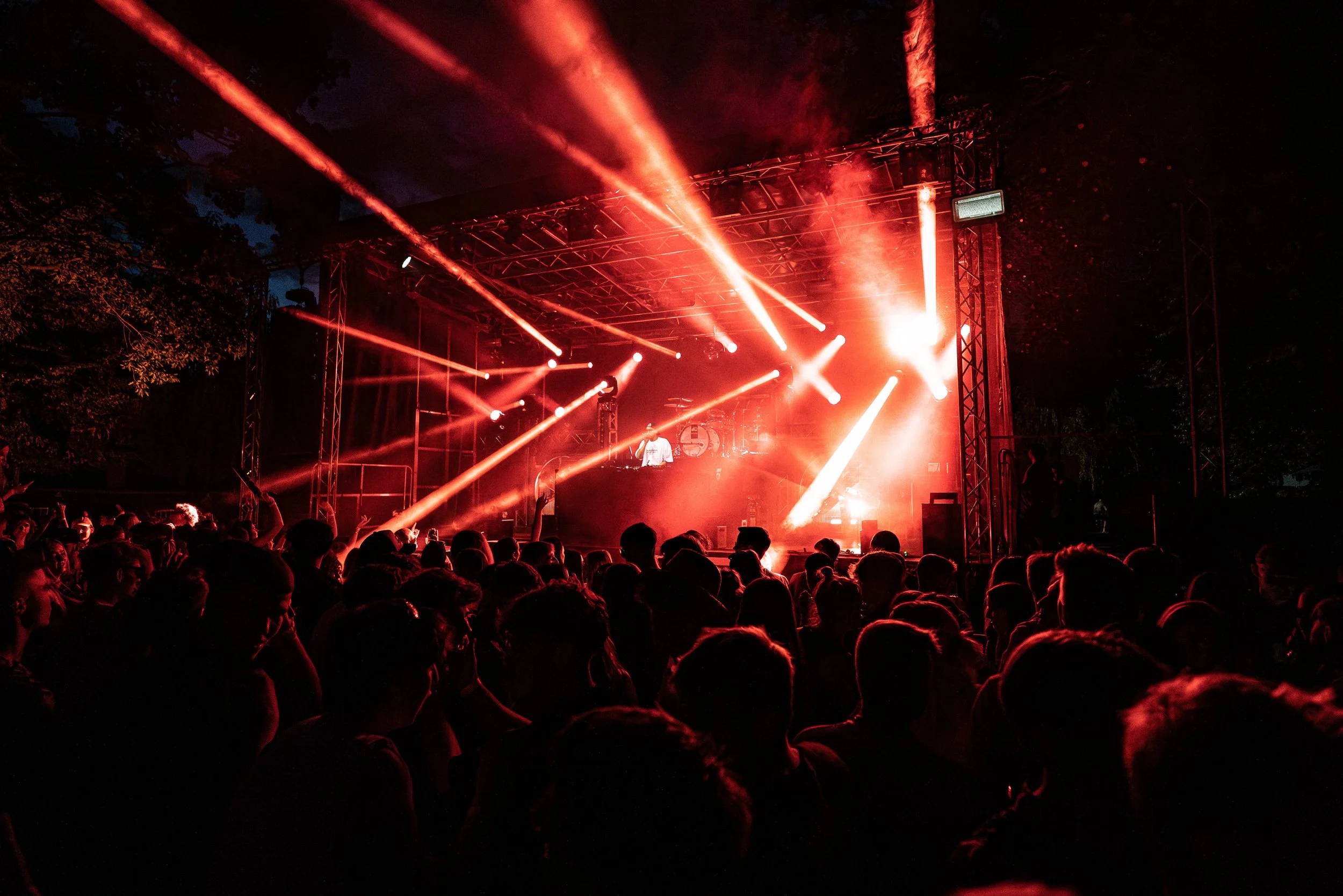 Bühne bei Nacht mit rotem Lichteffekt, ein DJ steht hinter einem Mischpult, vor der Bühne ein Publikum, das das Konzert anscheinend genießt.