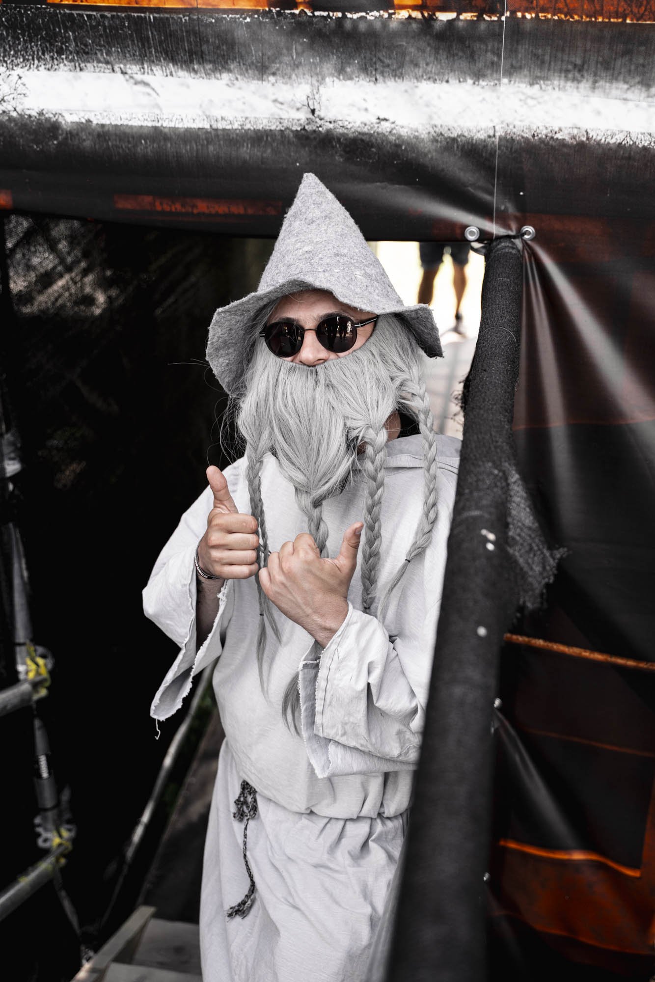 Person in Fantasiekostüm mit grauem Hut, weißem Bart und Brille, die einen Daumen hoch zeigt, versteckt sich hinter schwarzer Folie bei einem Fest.